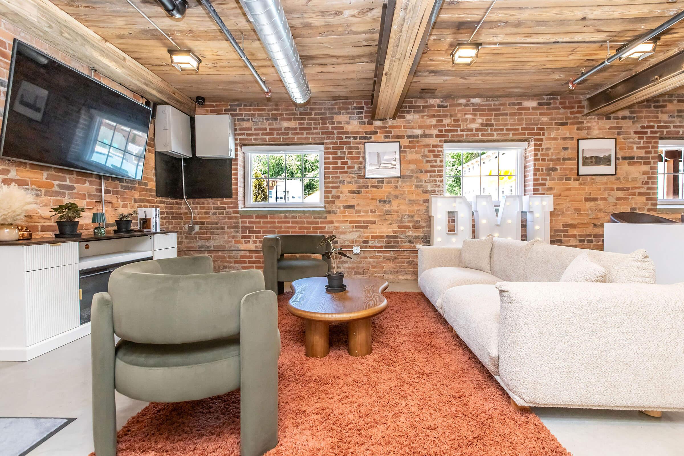 A modern living area featuring exposed brick walls, a wooden ceiling, and large windows. The space includes a plush beige sofa, stylish green armchairs, a wooden coffee table, and decorative items. A large TV is mounted on the wall, and illuminated letters spell "DMT" near the window. A warm rug adds coziness to the room.