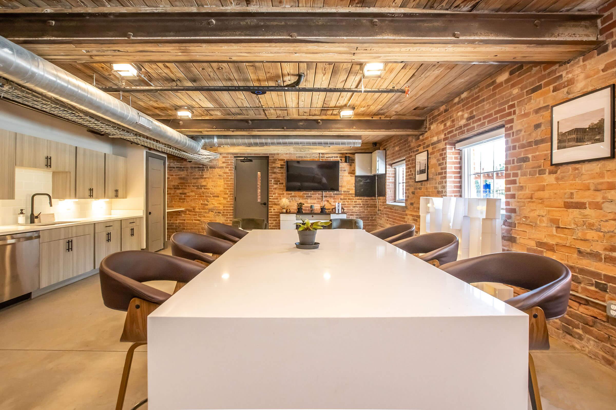 Modern conference room featuring a large white table surrounded by brown chairs, set in a stylish space with exposed brick walls and wooden beams. The room includes a kitchenette, a television mounted on the wall, and large windows providing natural light. A small potted plant sits in the center of the table.