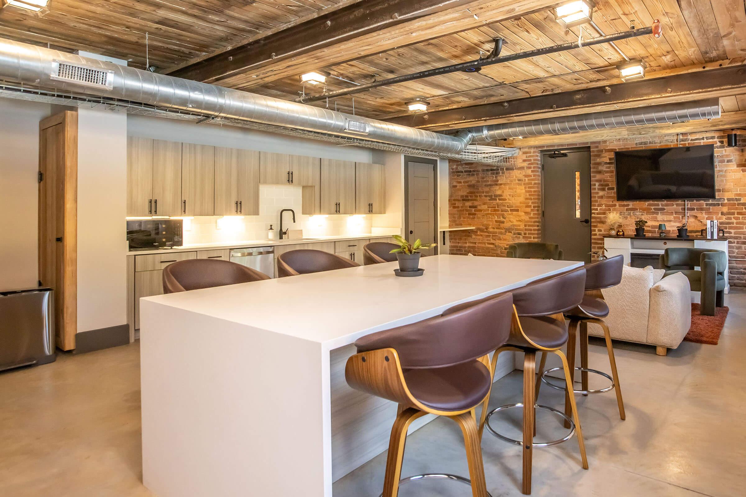 Modern kitchen and living area featuring a large white island with brown bar stools, sleek cabinetry, and stainless steel appliances. Exposed brick walls and wooden beams add character. A comfortable seating area with a TV is visible in the background. Natural light enhances the contemporary design.