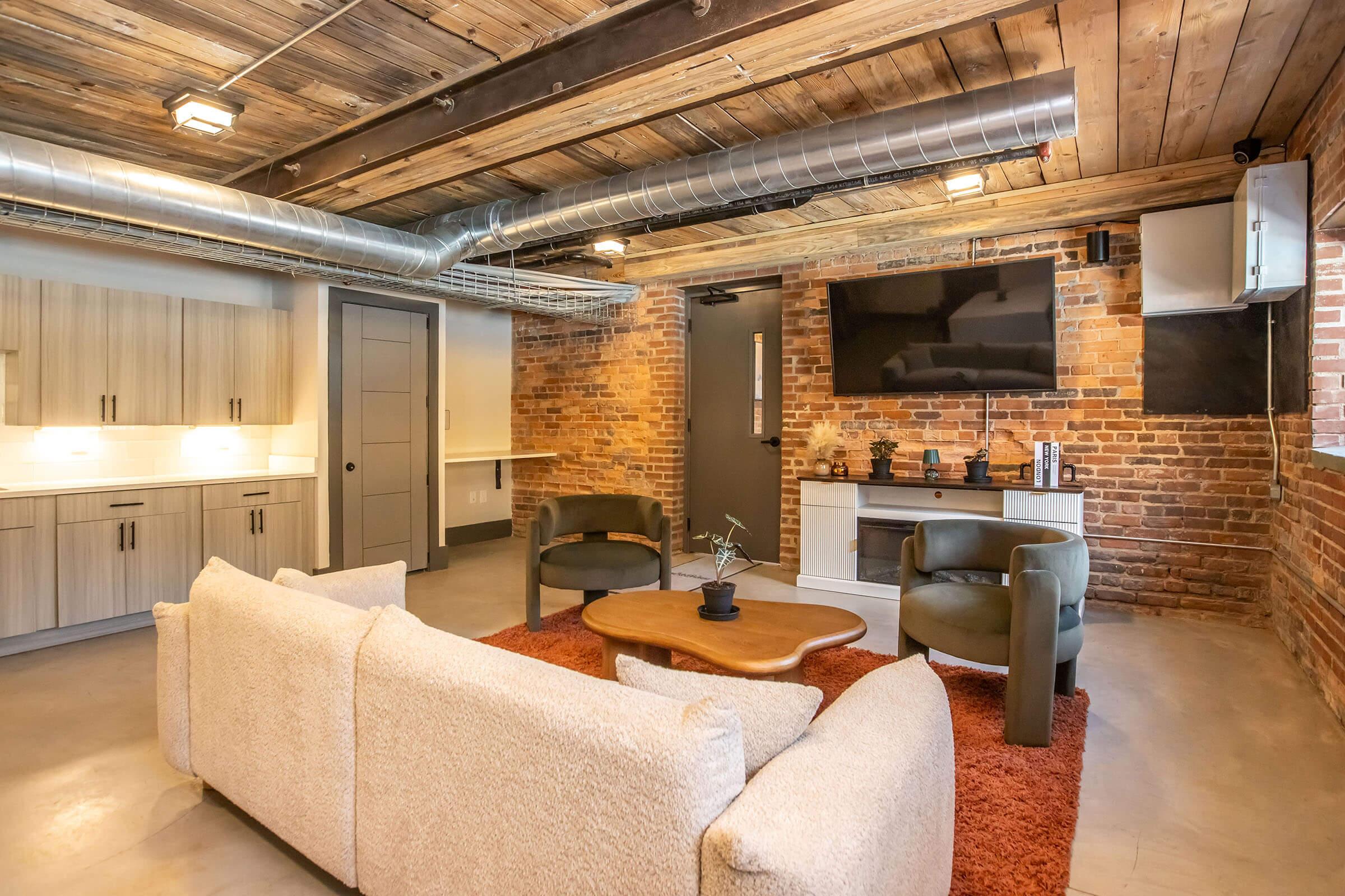 A modern, stylish living space featuring a cozy beige sofa, two green accent chairs, and a wooden coffee table on a vibrant orange rug. Exposed brick walls and wooden beams create a warm ambiance. A flat-screen TV is mounted on the wall, and there are sleek cabinets in the background, enhancing the contemporary decor.