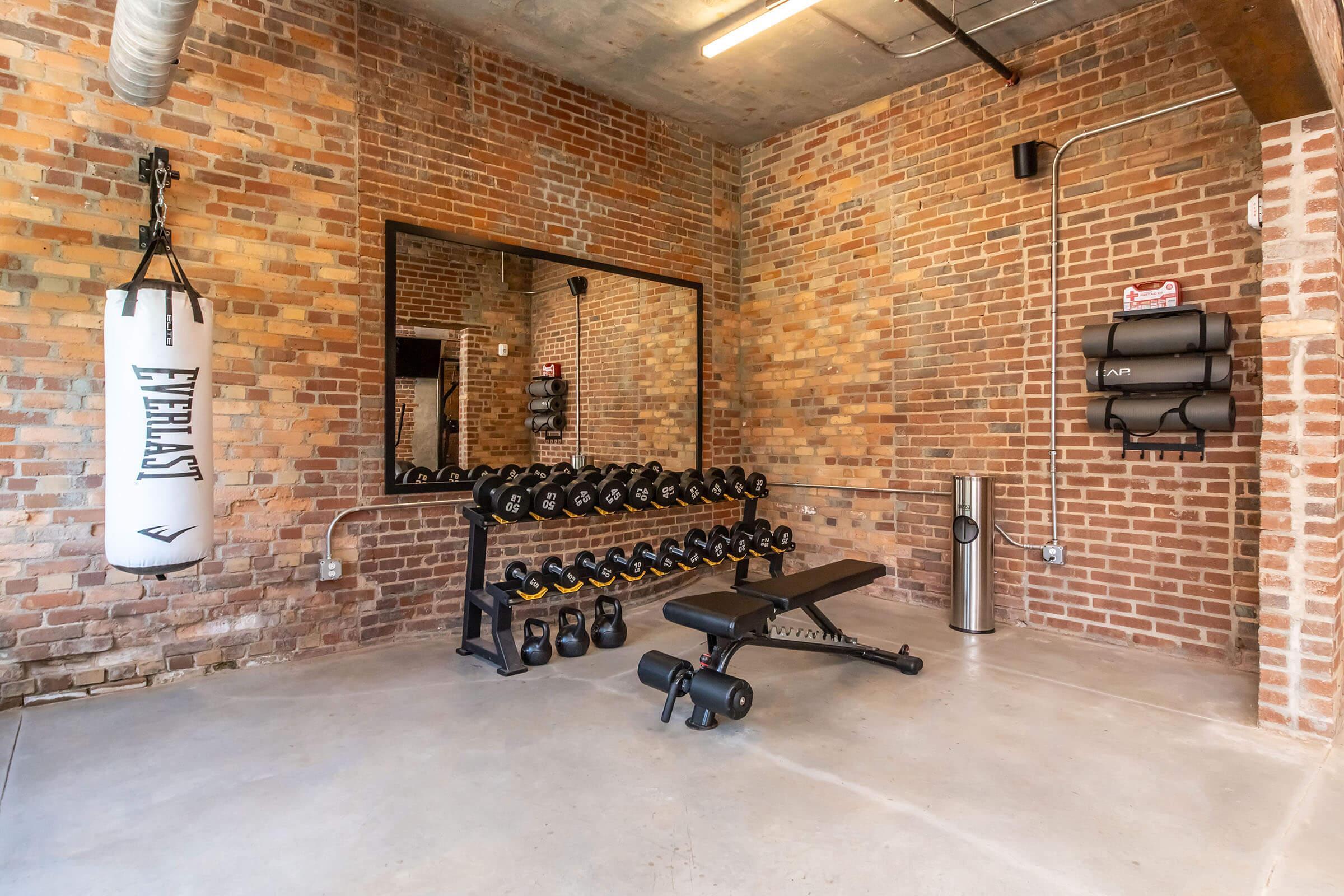 A bright, industrial-style home gym featuring exposed brick walls, a mirror, a set of dumbbells on a rack, a weight bench, a punching bag, and a water fountain. The space has a clean and modern aesthetic, with ample lighting and minimal decor.