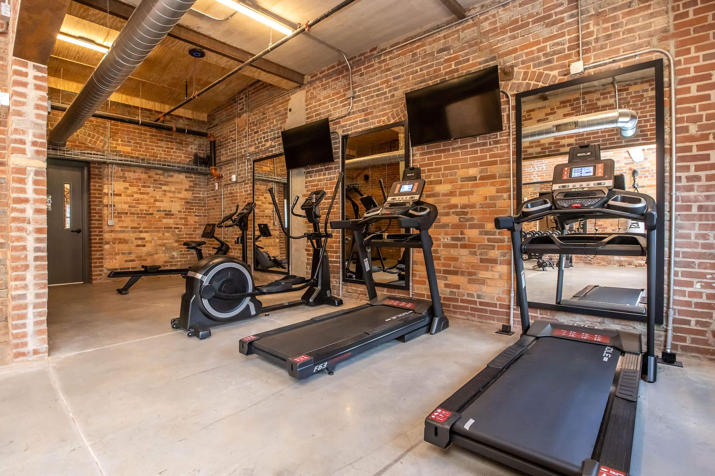 A modern gym interior featuring several exercise machines including treadmills and an elliptical, set against exposed brick walls. Large mirrors and two wall-mounted TVs are visible, creating a spacious and inviting workout environment. The floor is made of concrete, enhancing the industrial aesthetic.