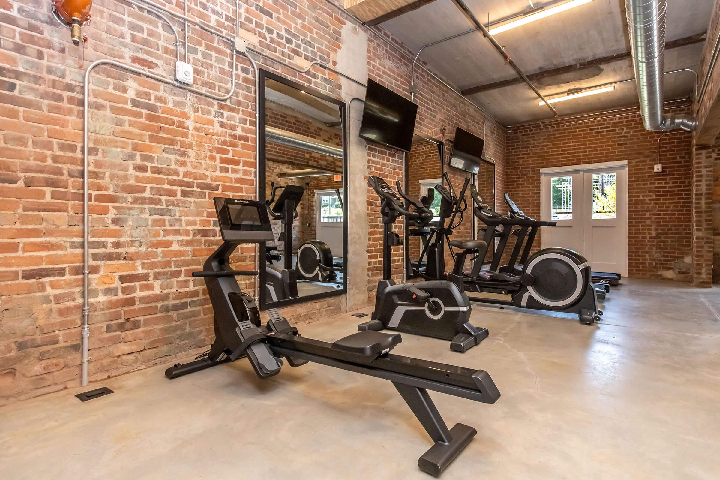 A modern gym interior featuring various exercise equipment, including a rowing machine and elliptical trainer. The space has exposed brick walls, large mirrors, and wall-mounted televisions, creating a bright and open workout environment. Large windows allow natural light to filter in.