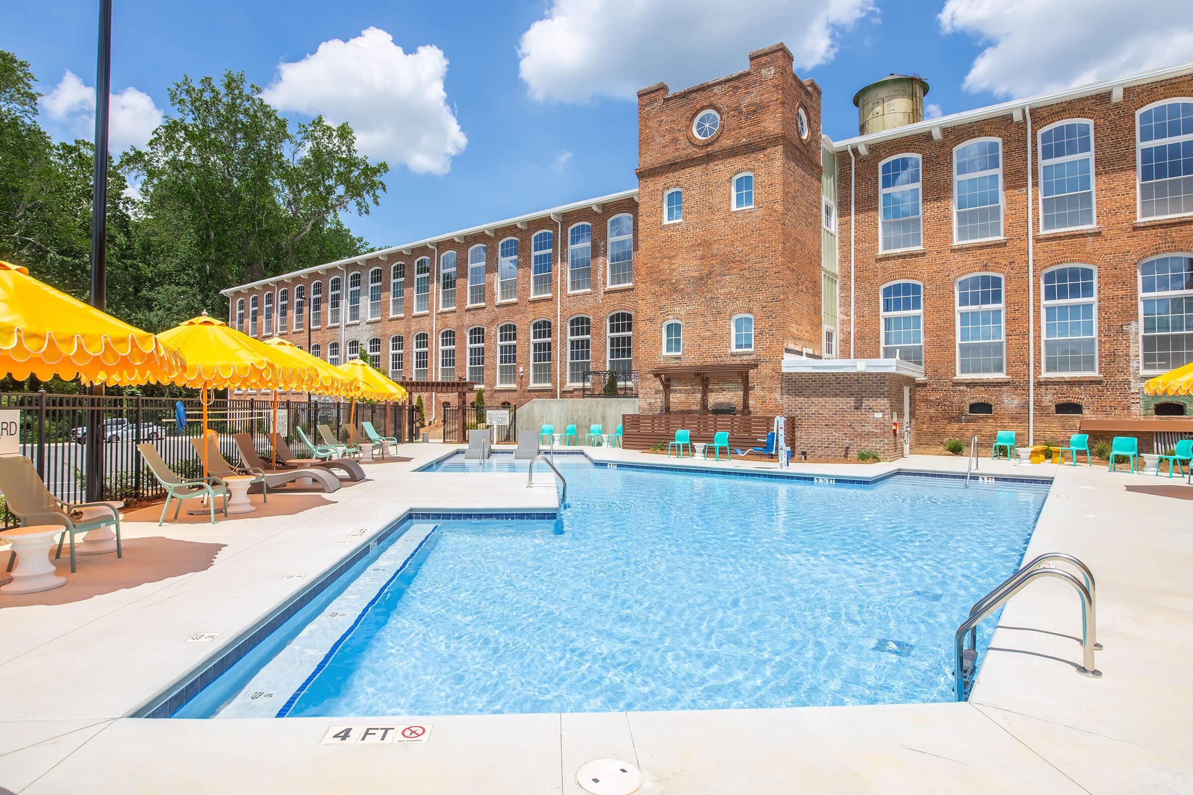 A bright, inviting outdoor pool area with clear blue water, surrounded by lounge chairs and yellow umbrellas. In the background, a historic red brick building with large windows and a clock tower adds character to the setting. Trees and a blue sky with fluffy clouds complete the scene.