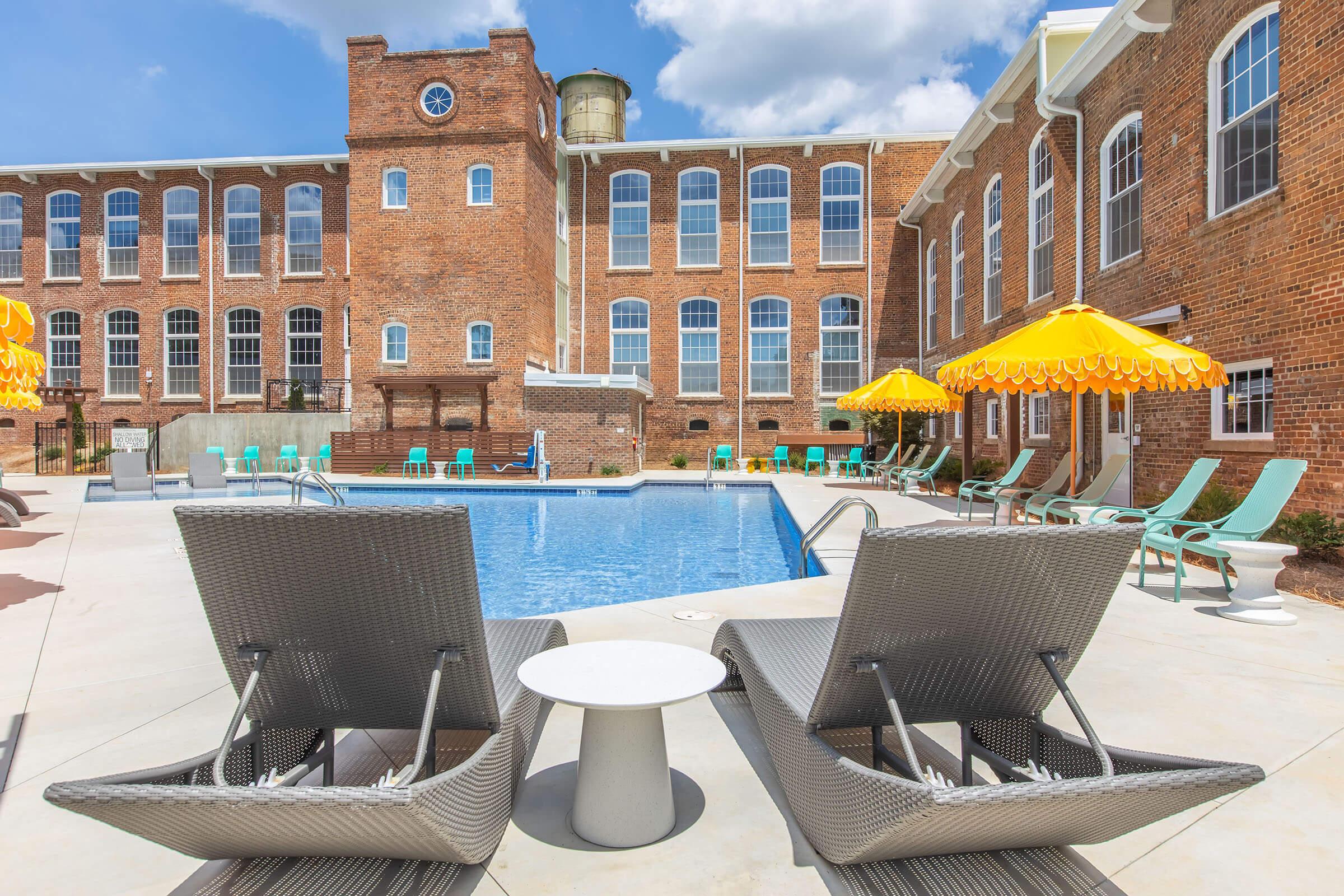 A pool area featuring a clear blue swimming pool surrounded by lounge chairs and yellow umbrellas. In the background, a brick building with large windows and a water tower is visible, complementing the inviting outdoor space. Bright skies enhance the overall summer atmosphere.