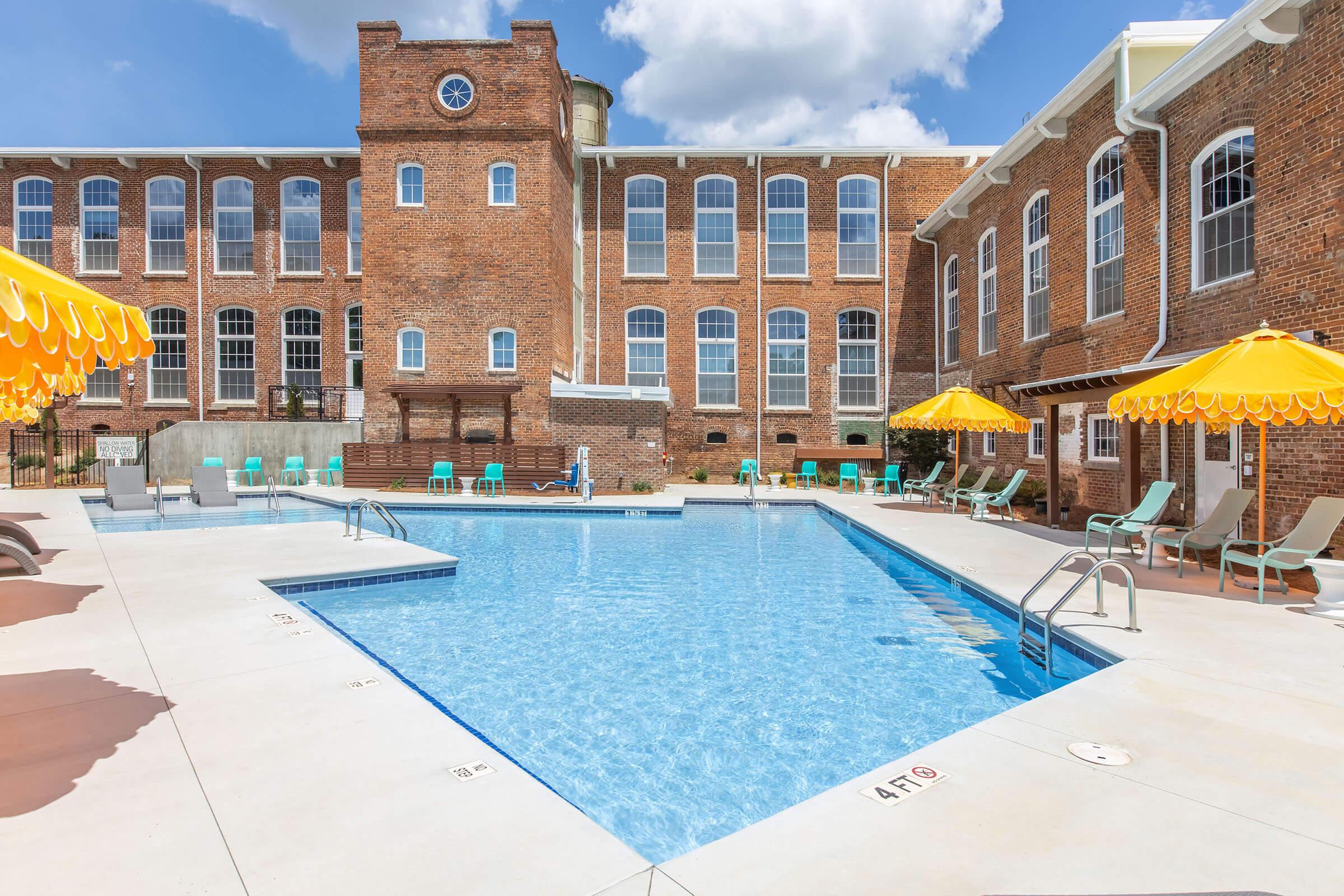 A bright outdoor swimming pool with lounge chairs and yellow umbrellas, surrounded by a brick building with large windows. The sky is partly cloudy, and the pool area is well-lit and inviting, perfect for relaxation or recreation.
