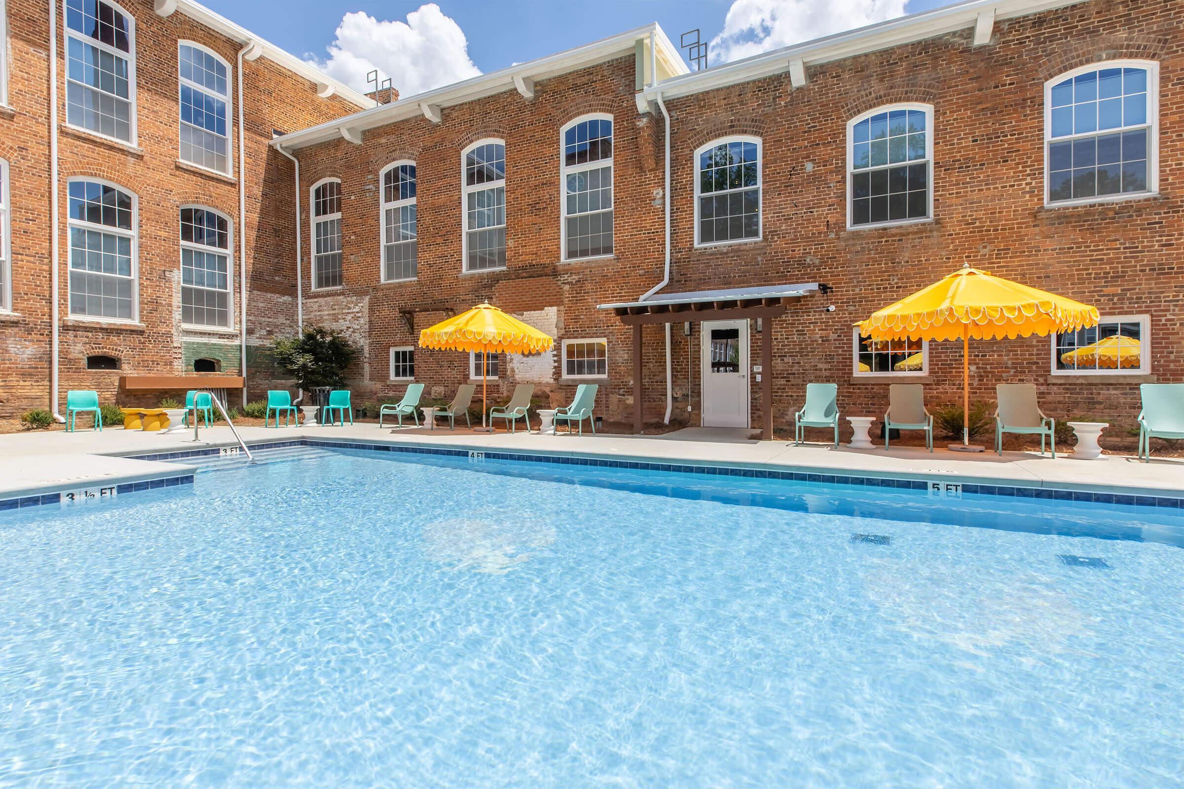 A clear blue swimming pool in front of a brick building with large windows. Poolside, there are yellow umbrellas shading green lounge chairs. The scene is well-lit, showcasing a relaxing outdoor area ideal for leisure and socializing.