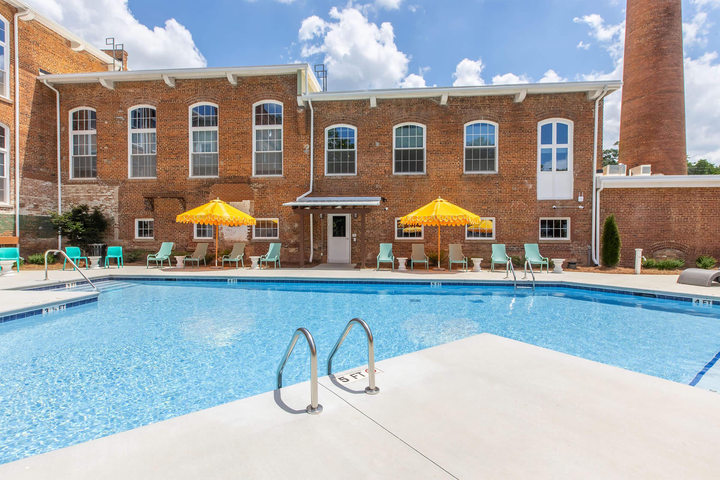 A bright swimming pool surrounded by lounge chairs and yellow umbrellas, with a historic brick building in the background. The sky is partly cloudy, and there is a large brick chimney visible. The pool area is well-maintained and inviting for relaxation and leisure activities.