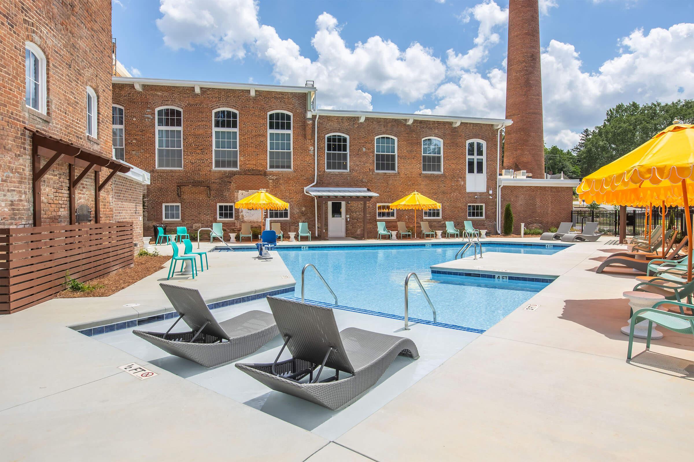 A sunny outdoor pool area featuring lounge chairs beside a swimming pool. Yellow umbrellas provide shade, with brick buildings and a tall chimney in the background. The scene is bright and inviting, perfect for relaxation.