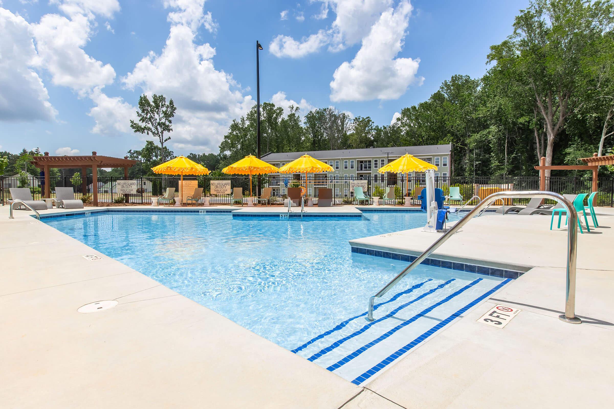 A clear swimming pool surrounded by lounge chairs and yellow umbrellas, with a grassy area and trees in the background. The pool features a shallow area with steps for entry, and there are sunlit clouds in the blue sky above.
