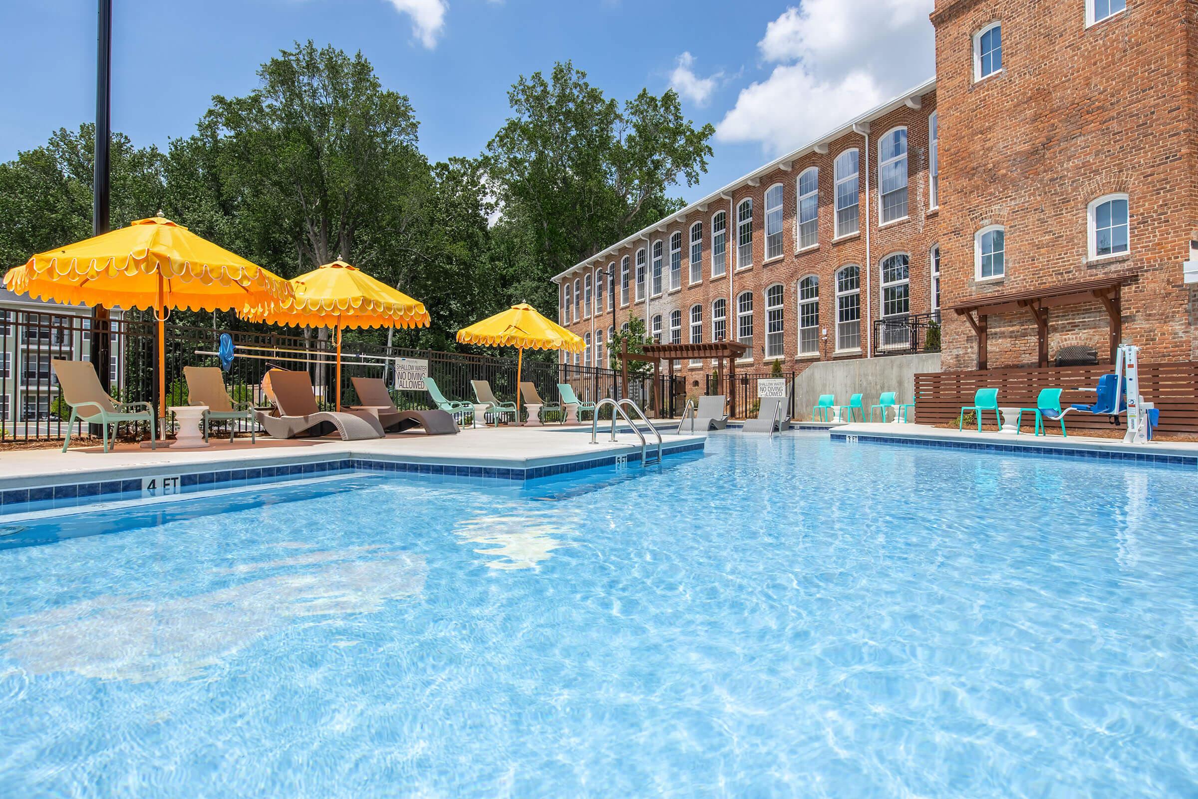 A sunny outdoor pool area featuring clear blue water, lounge chairs, and yellow umbrellas. The brick building in the background has large windows, surrounded by green trees and a fence. The scene creates a relaxing atmosphere perfect for leisure and enjoyment.