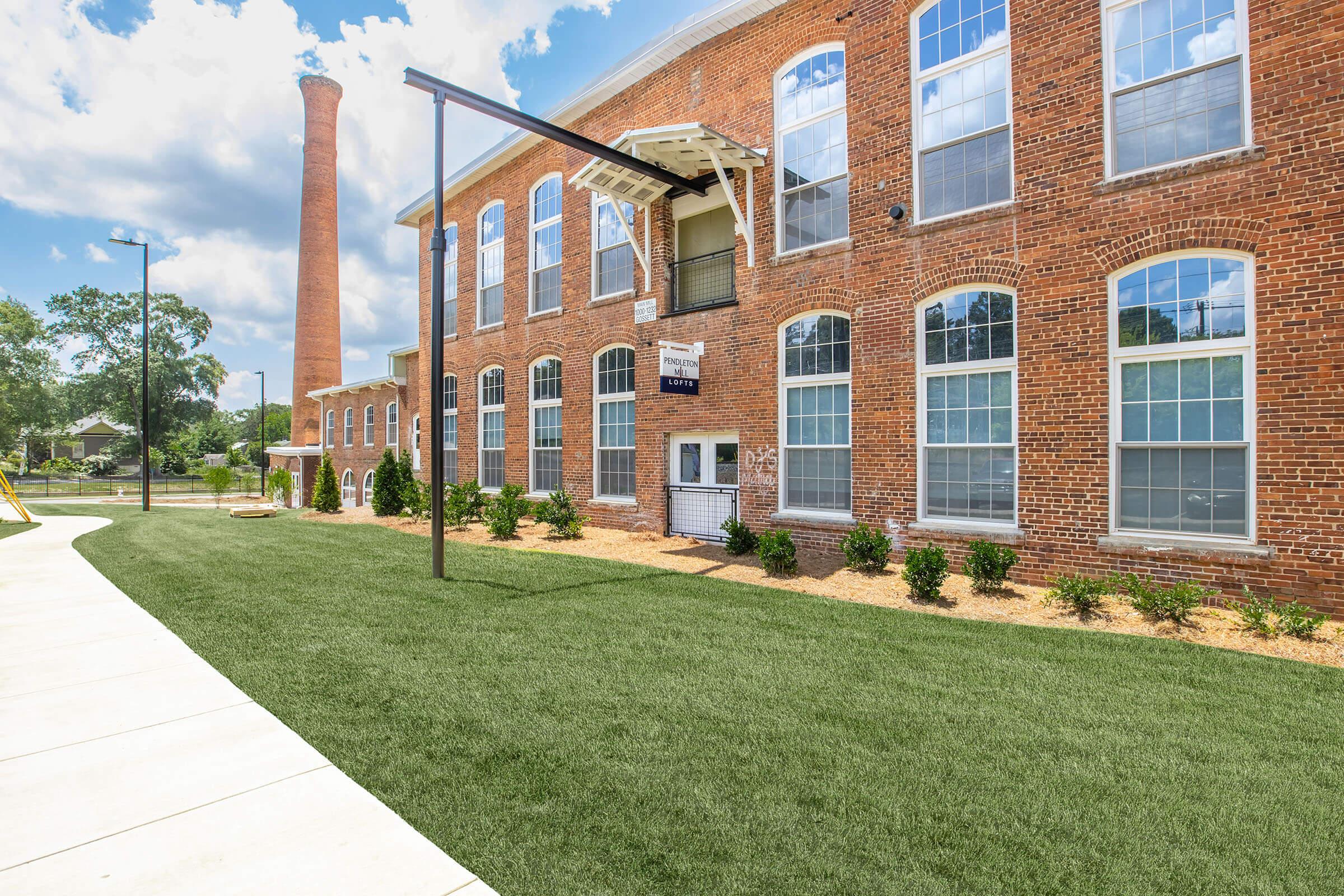 A red brick building with large windows, featuring a white balcony and an entrance with a sign. Nearby, there is a tall brick chimney and well-maintained green grass. The sky is partly cloudy, creating a bright atmosphere in the outdoor setting.