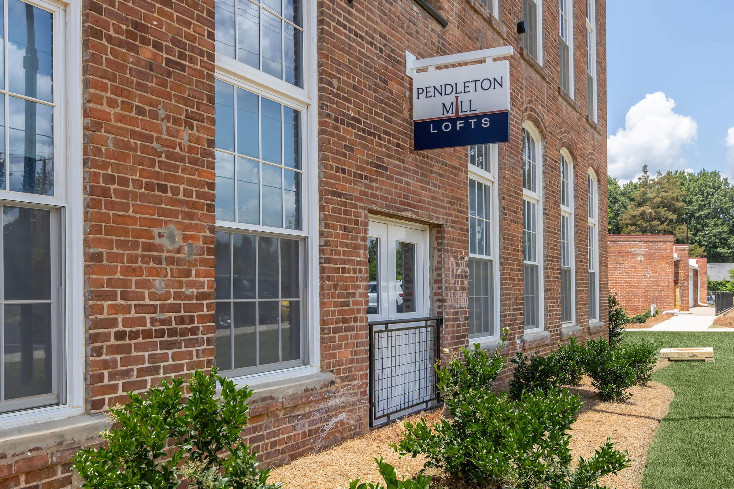 Exterior of Pendleton Mill Lofts, a multi-story brick building featuring large windows and a prominent sign. The ground floor has a small entrance with a decorative gate. Lush greenery and freshly laid gravel surround the building, under a clear blue sky with a few clouds in the background.