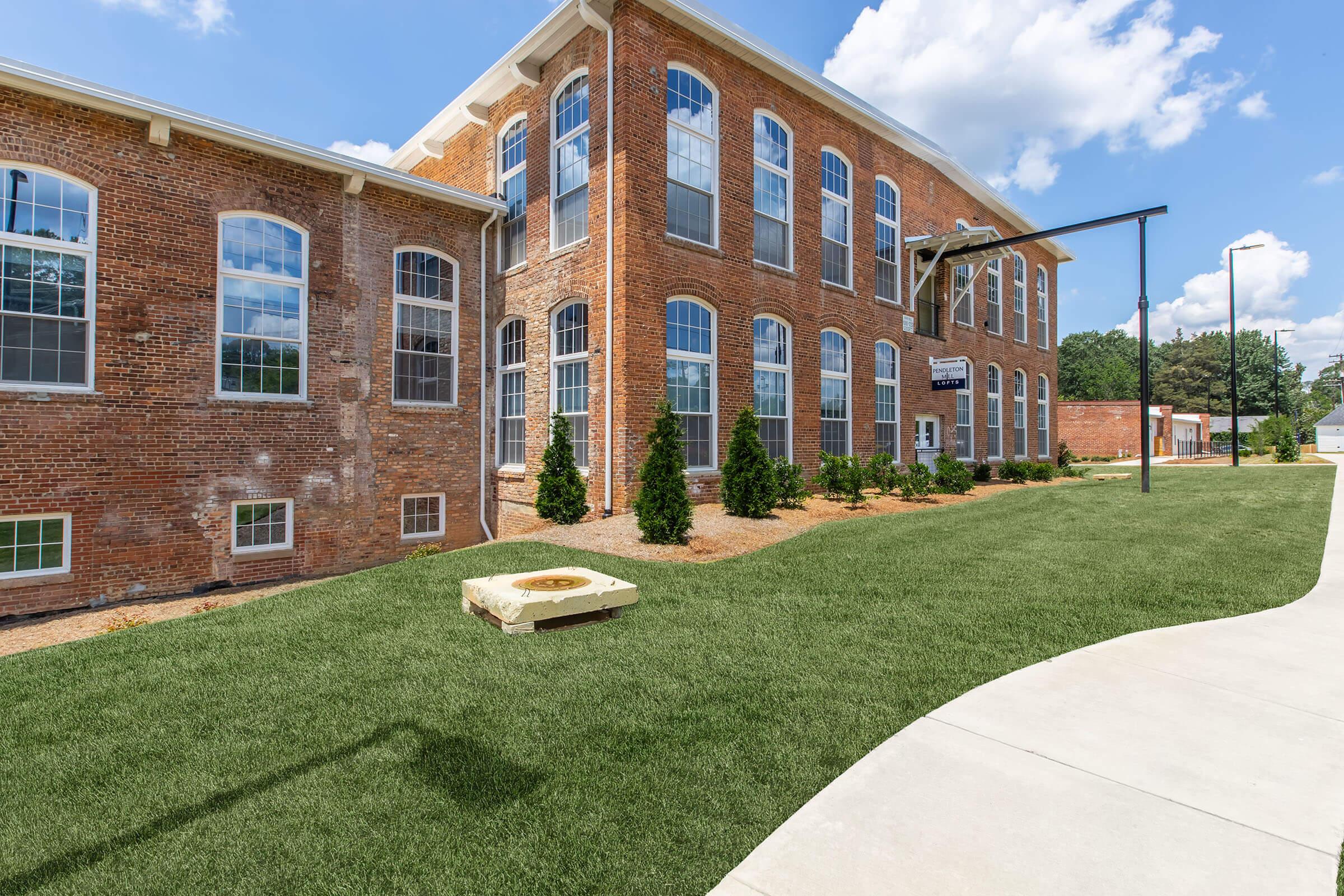 Exterior view of a red brick building with large windows, surrounded by landscaped greenery. A small paved pathway leads up to the building, which features a sign visible on the side. The sky is partly cloudy, enhancing the bright and welcoming atmosphere.