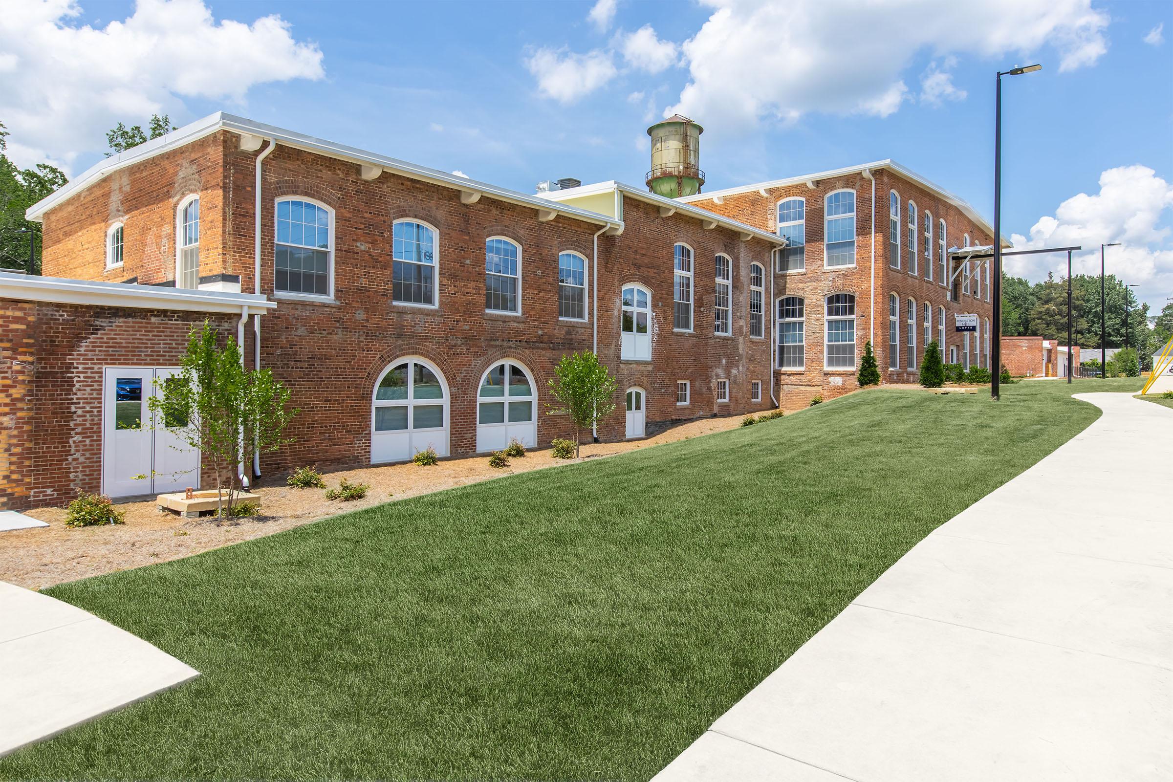 A brick building with large arched windows, surrounded by a green lawn. The structure features a water tower and is situated in a bright, sunny environment with a blue sky and fluffy clouds. There are pathways leading up to the building and landscaped areas with small trees.