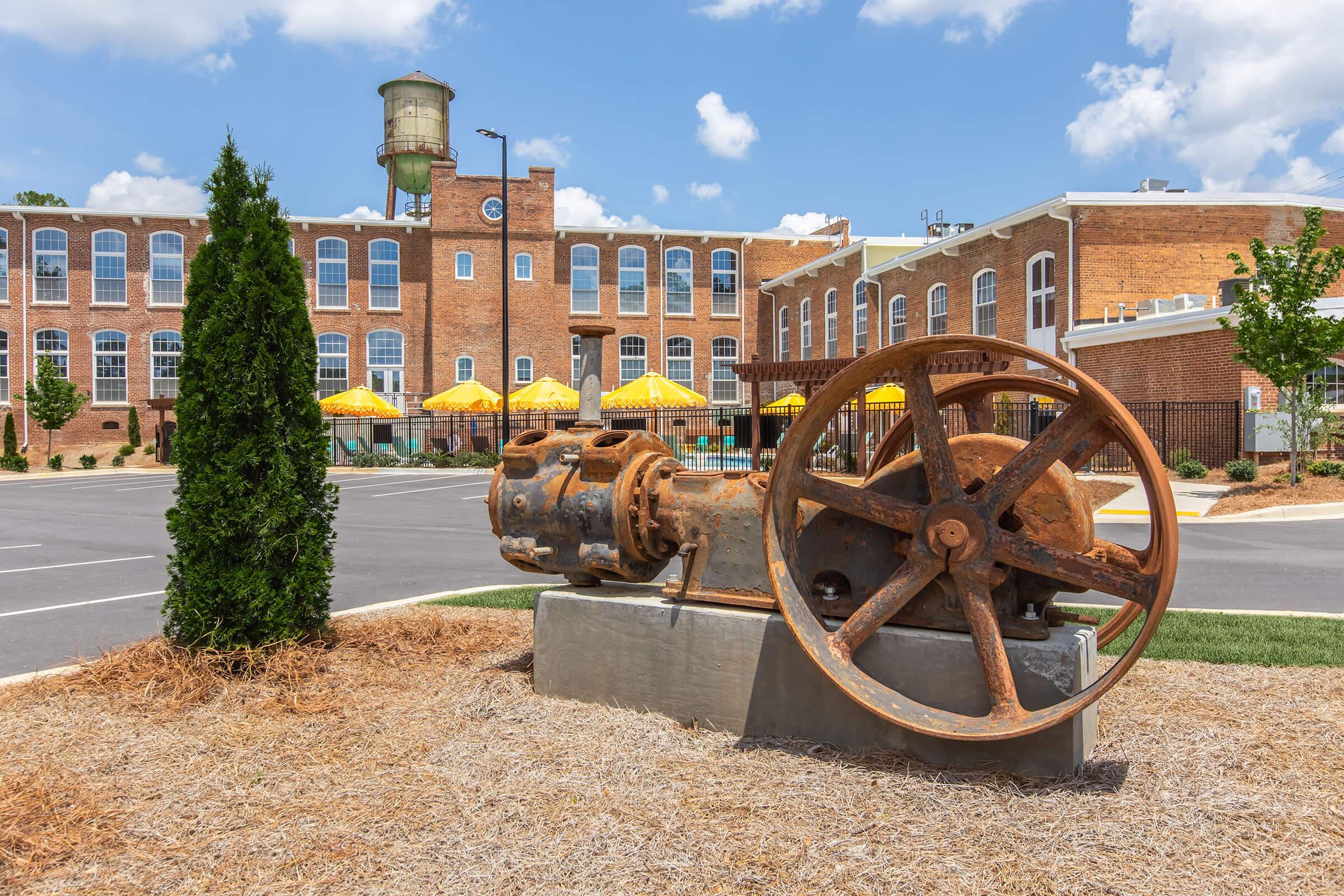 A rusty, large gear sits on a stone pedestal in the foreground, with a picturesque building in the background. The building features large windows and is part of a renovated industrial complex. Yellow umbrellas are visible, suggesting outdoor seating. A water tower is also present against a blue sky with fluffy clouds.