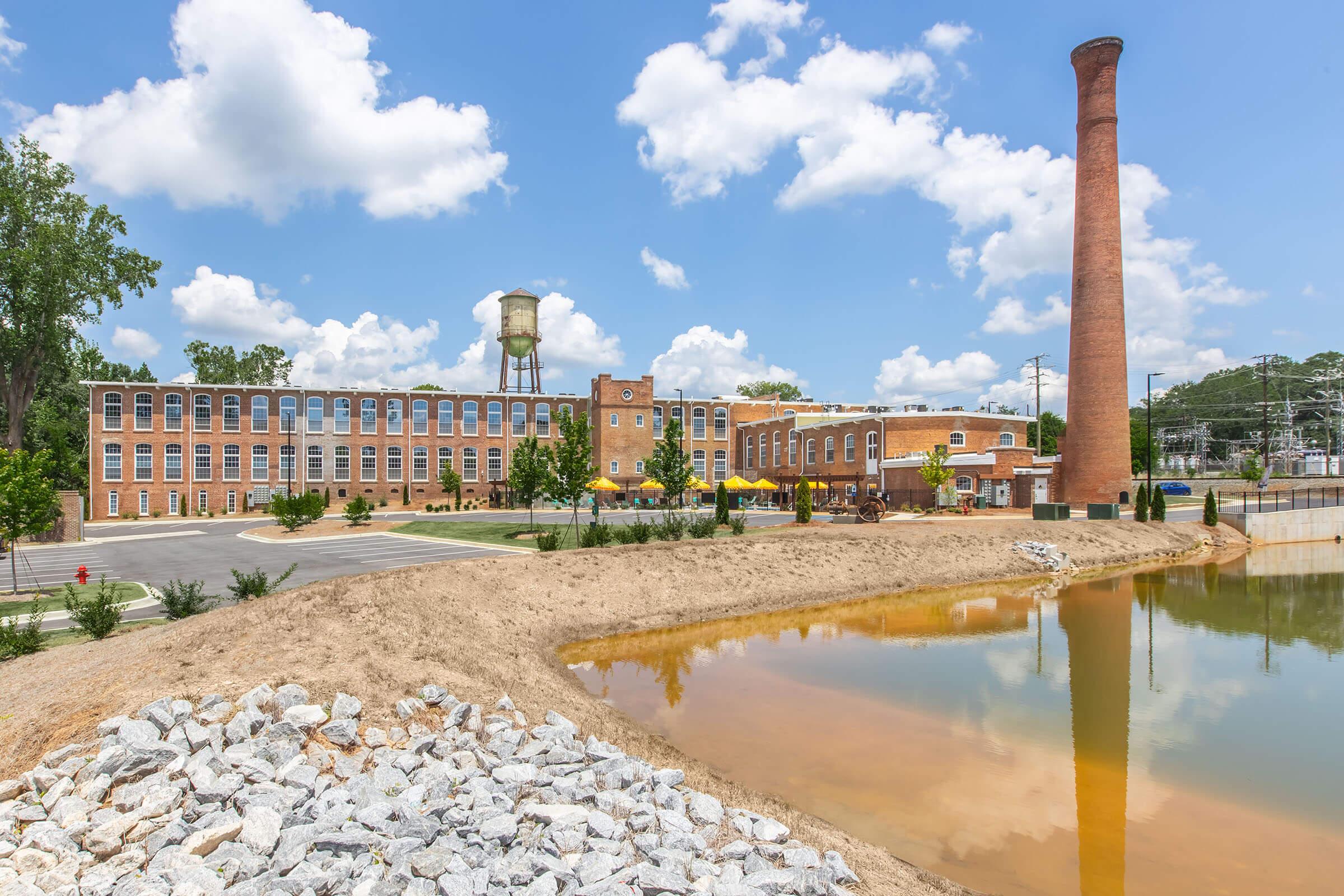 Historic industrial building with a tall brick chimney, water tower, and surrounding landscaping. Reflections in a pond in the foreground, with rocky embankments. The scene is set under a bright blue sky dotted with fluffy white clouds, showcasing a blend of nature and restored architecture.