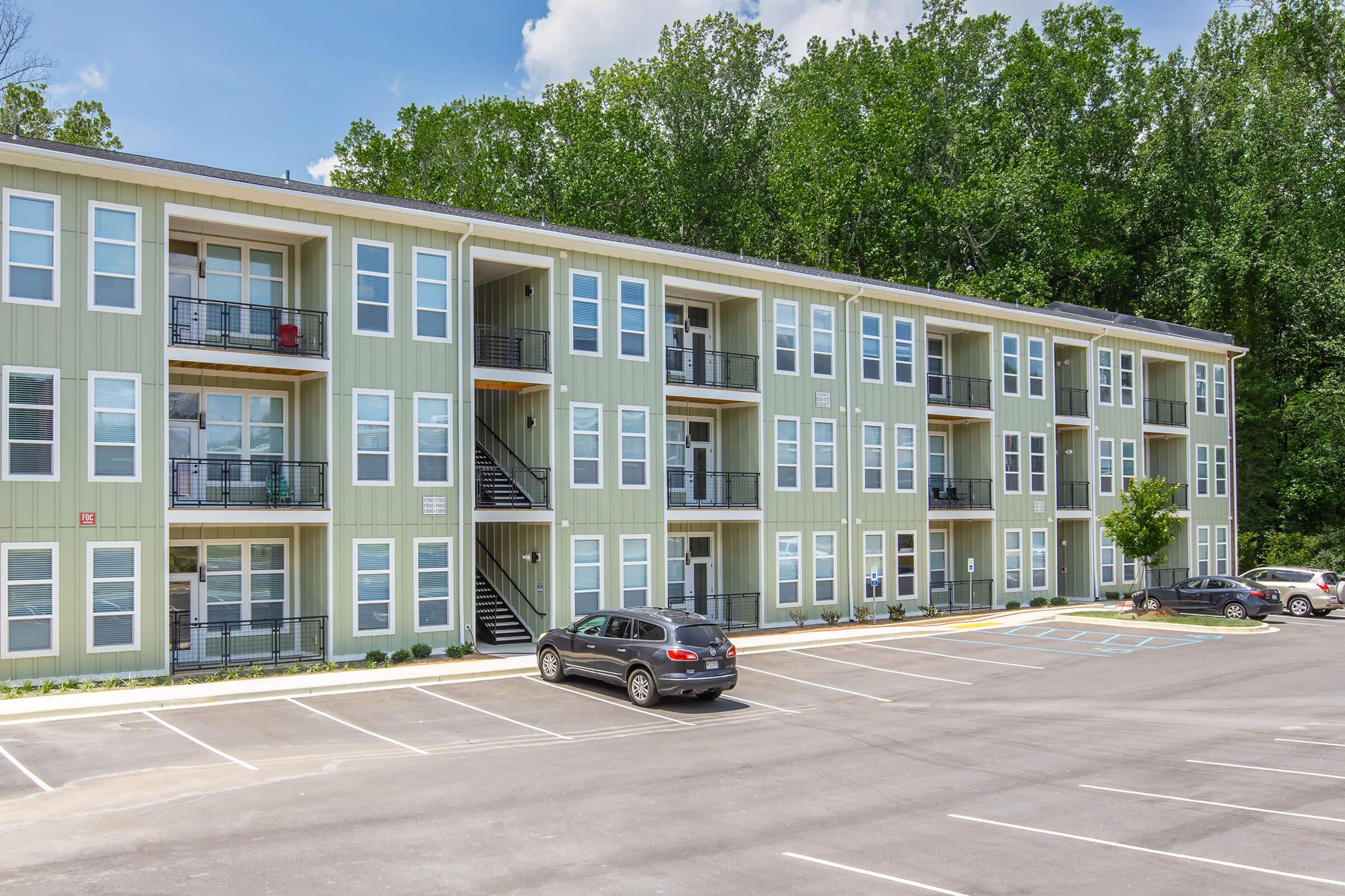 A multi-story apartment building with a green exterior, featuring multiple balconies and large windows. A parking lot is in front, with several cars parked. Lush trees provide a natural backdrop, and the sky is partly cloudy.