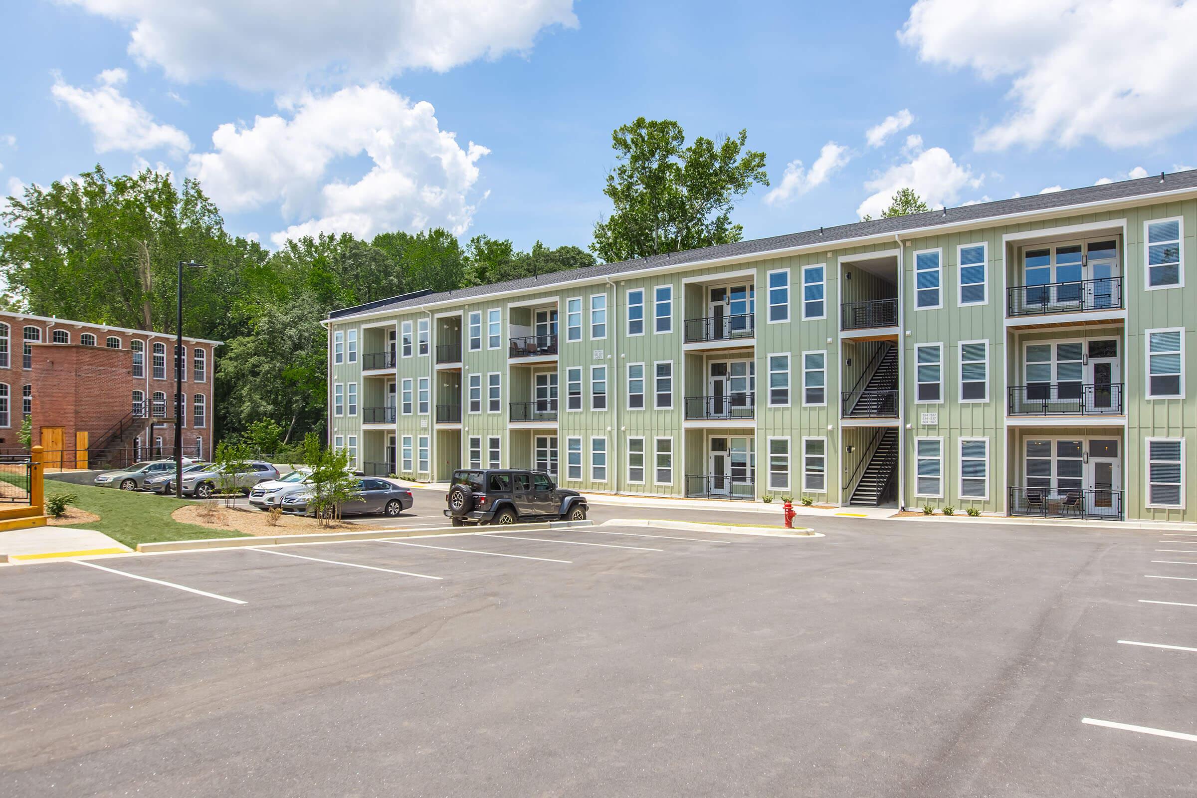A modern, multi-story apartment building with green siding and multiple balconies. The scene features a paved parking area with several vehicles and a background of trees under a partly cloudy sky. Nearby, there is another building visible, adding to the residential setting.