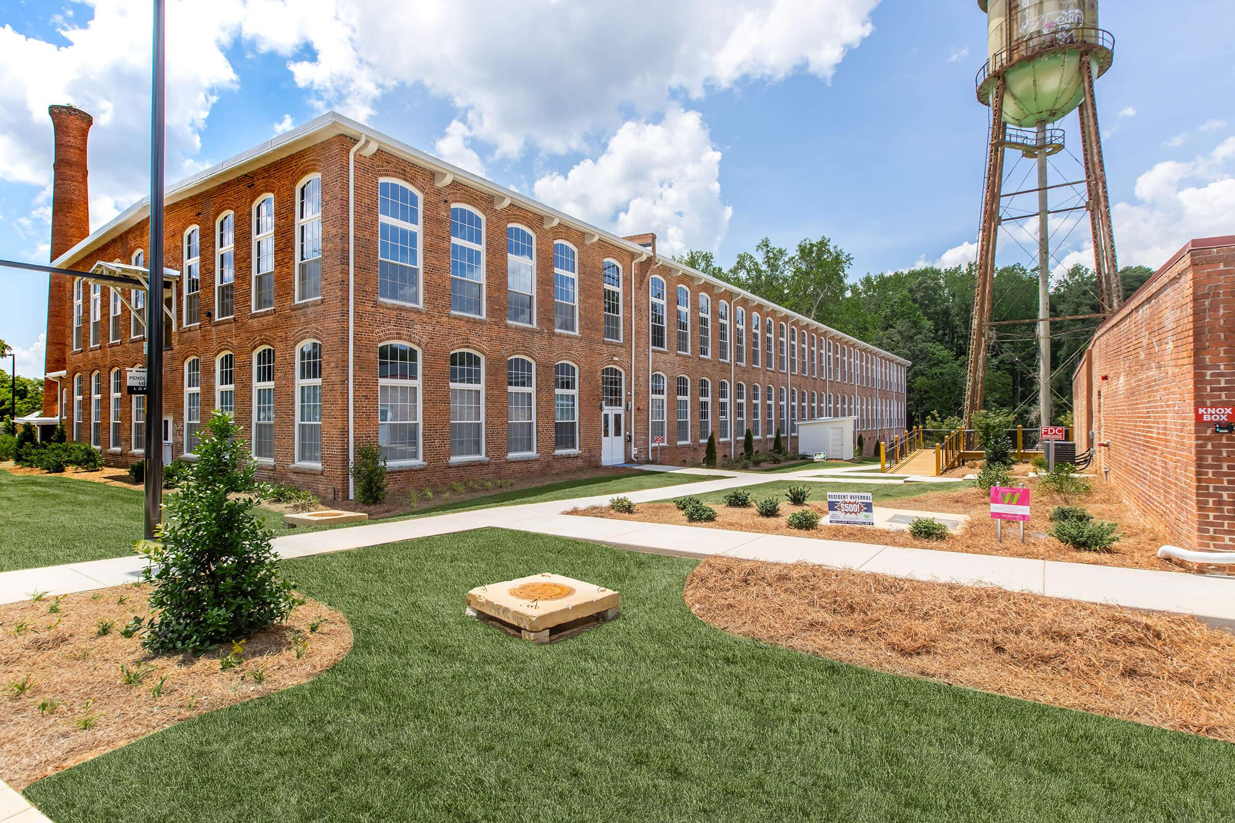 A historic brick factory building with large windows, situated among green landscaping. A water tower is visible in the background, and the scene features a well-maintained pathway and grass area. Bright blue sky with scattered clouds enhances the industrial charm of the setting.