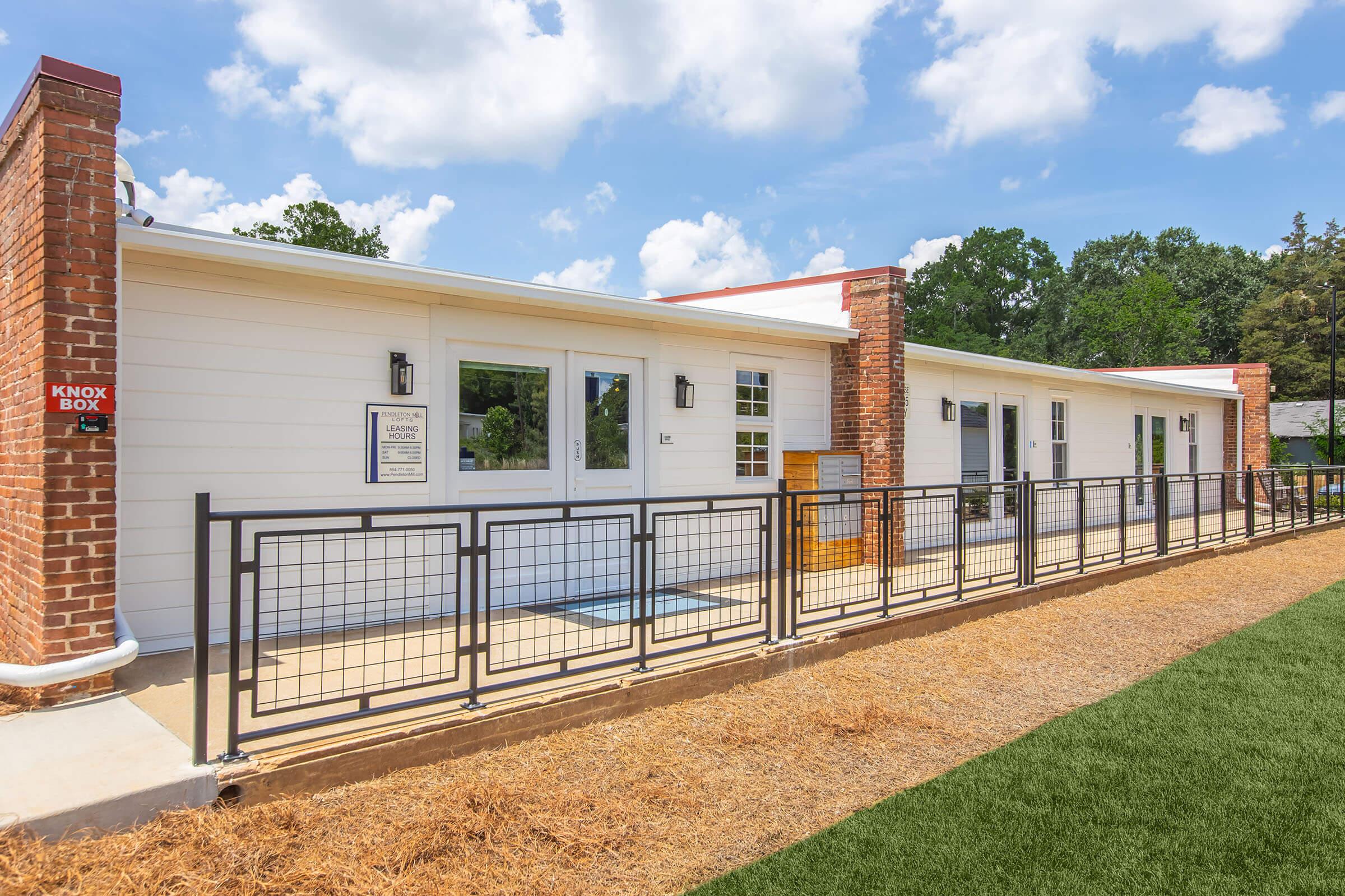 A modern single-story building with a white exterior and brick accents, featuring several windows and doors. The pathway is bordered by a metal railing, and there's a grassy area in front. A Knox Box is visible on the side of the building. The sky is partly cloudy, adding a bright atmosphere to the scene.