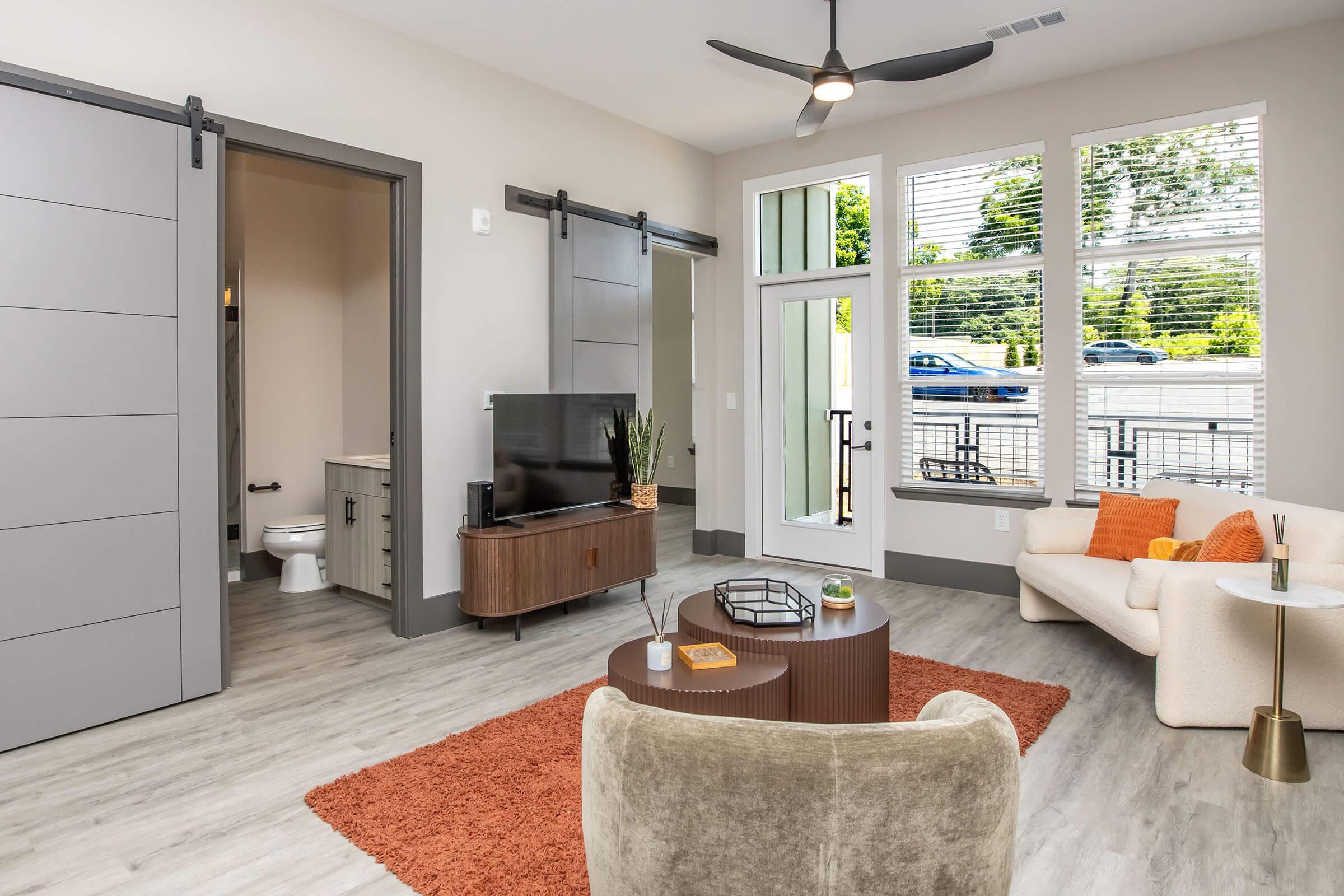 Modern living room featuring a light-colored sofa, a round coffee table, and a plush chair. A large TV is on a low console table. The room has bright natural light from large windows, with sliding doors leading to an outside area. The floor is a light wood finish, and an area rug adds warmth to the space.