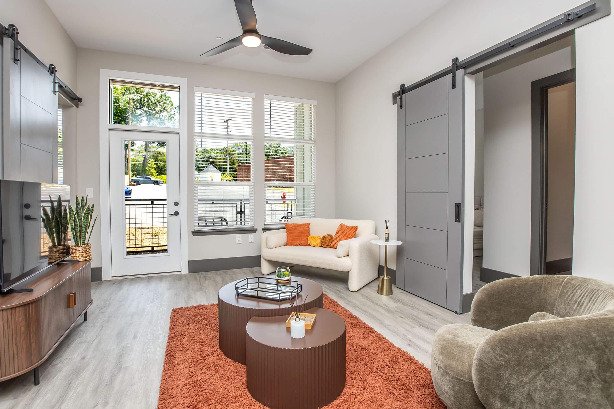A modern living room featuring a light-colored sofa with orange accents, two round coffee tables, a textured orange rug, and large windows with blinds. There is a flat-screen TV on a wooden media console and a cozy armchair. A sliding door leads to another room, enhancing the contemporary aesthetic.