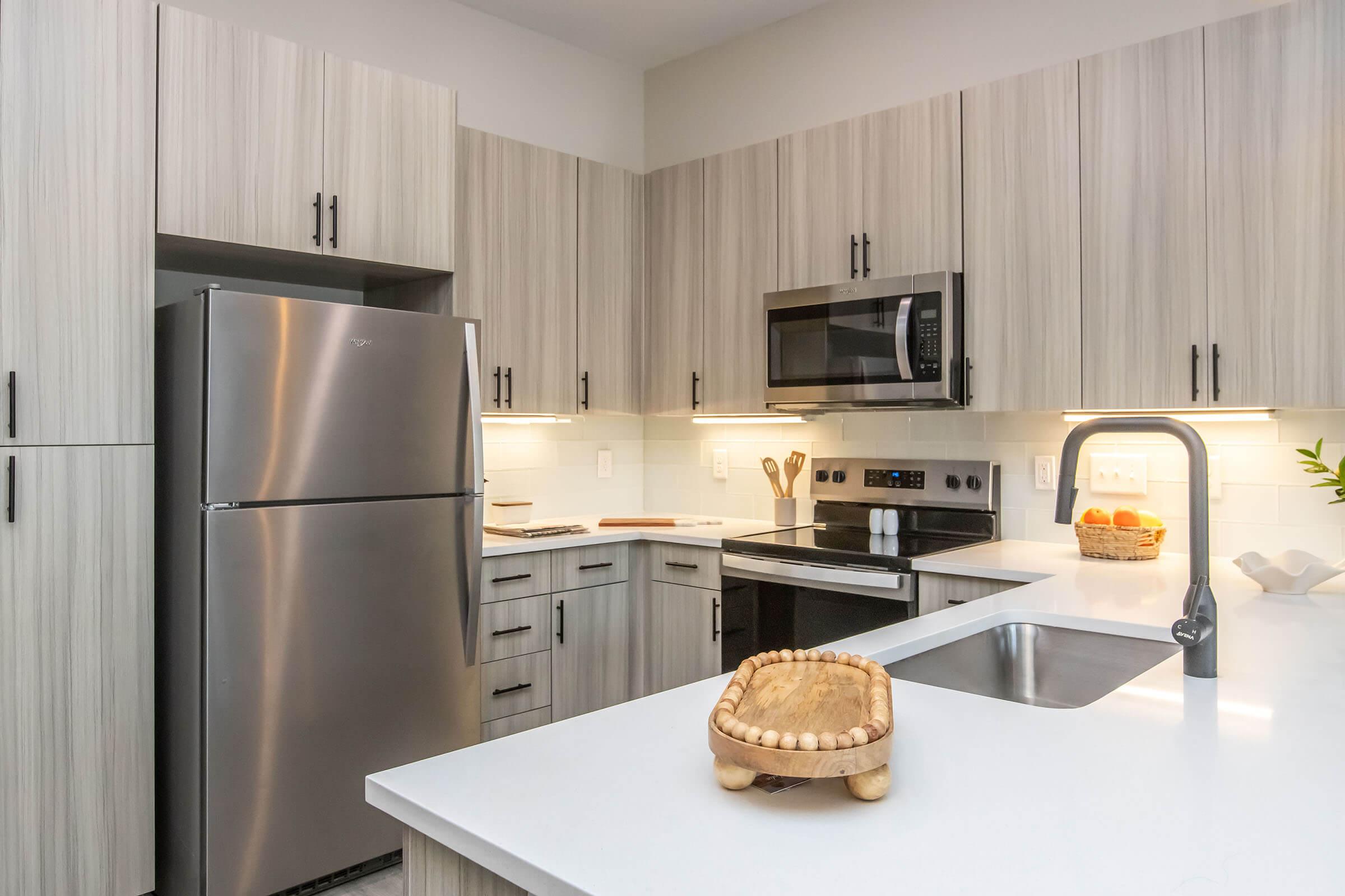 Modern kitchen featuring light wood cabinetry, a stainless steel refrigerator, microwave, and stove. A white countertop with a wooden bowl is in the foreground, while subtle lighting highlights the sleek design. The overall aesthetic is contemporary and functional.