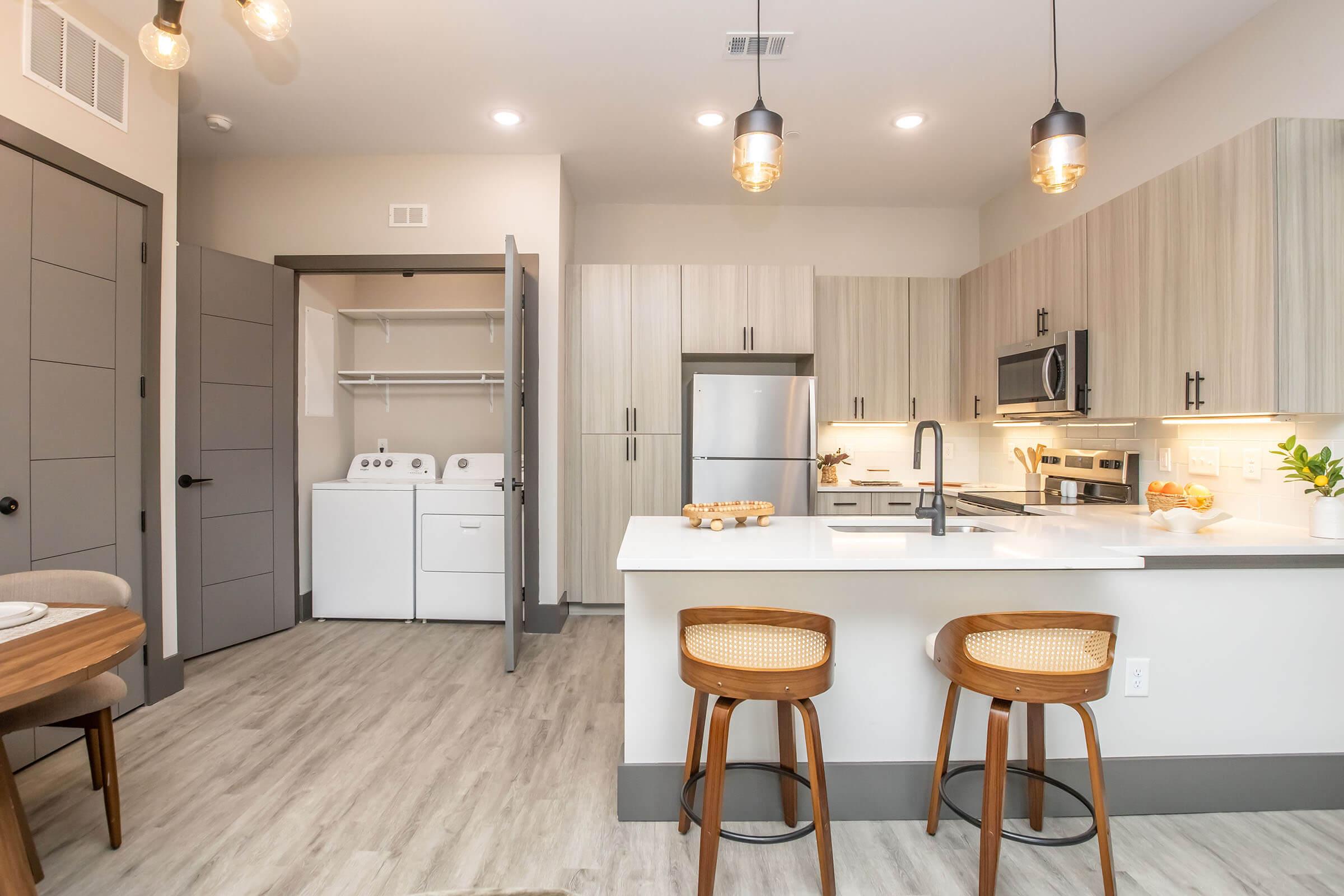 Modern kitchen with light wood cabinetry, stainless steel appliances, and a large island with seating. A dining area is visible with a round table. In the background, a laundry area is shown. The space is well-lit with pendant lighting and features a neutral color palette.