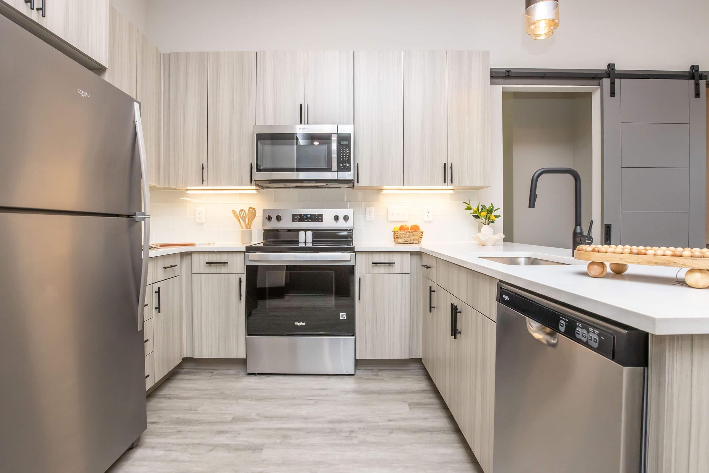 Modern kitchen featuring light wood cabinetry, stainless steel appliances including a refrigerator, microwave, and stove. A black faucet sits over a white sink, and natural light comes from an adjacent room. The countertop has kitchen utensils and a bowl of fruit, with a rolling pin placed beside the sink.