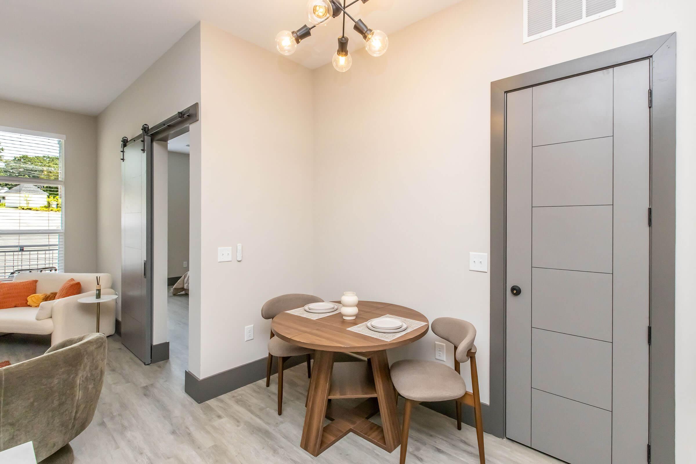 A cozy dining area featuring a round wooden table with two place settings, accompanied by two upholstered chairs. The wall behind has a modern sliding door, and a light fixture with exposed bulbs hangs from the ceiling. The walls are painted a soft beige, creating a warm and inviting atmosphere.