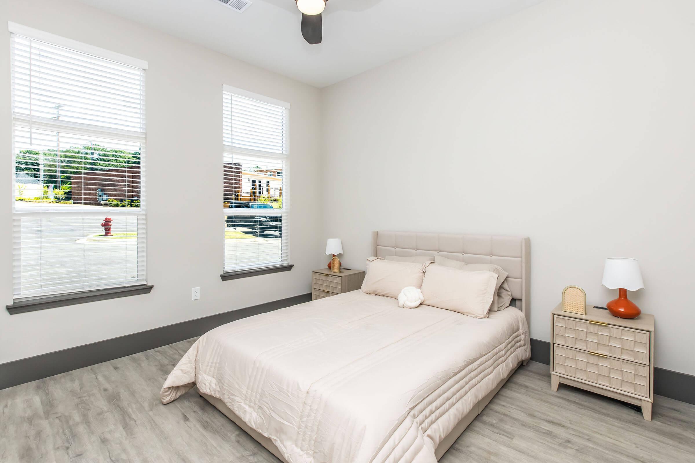 A bright and minimalist bedroom featuring a queen-sized bed with a light-colored quilt. Two bedside tables with lamps are positioned on either side. The walls are painted a soft beige, and there are two windows allowing natural light to illuminate the space, with a view of an outdoor area.