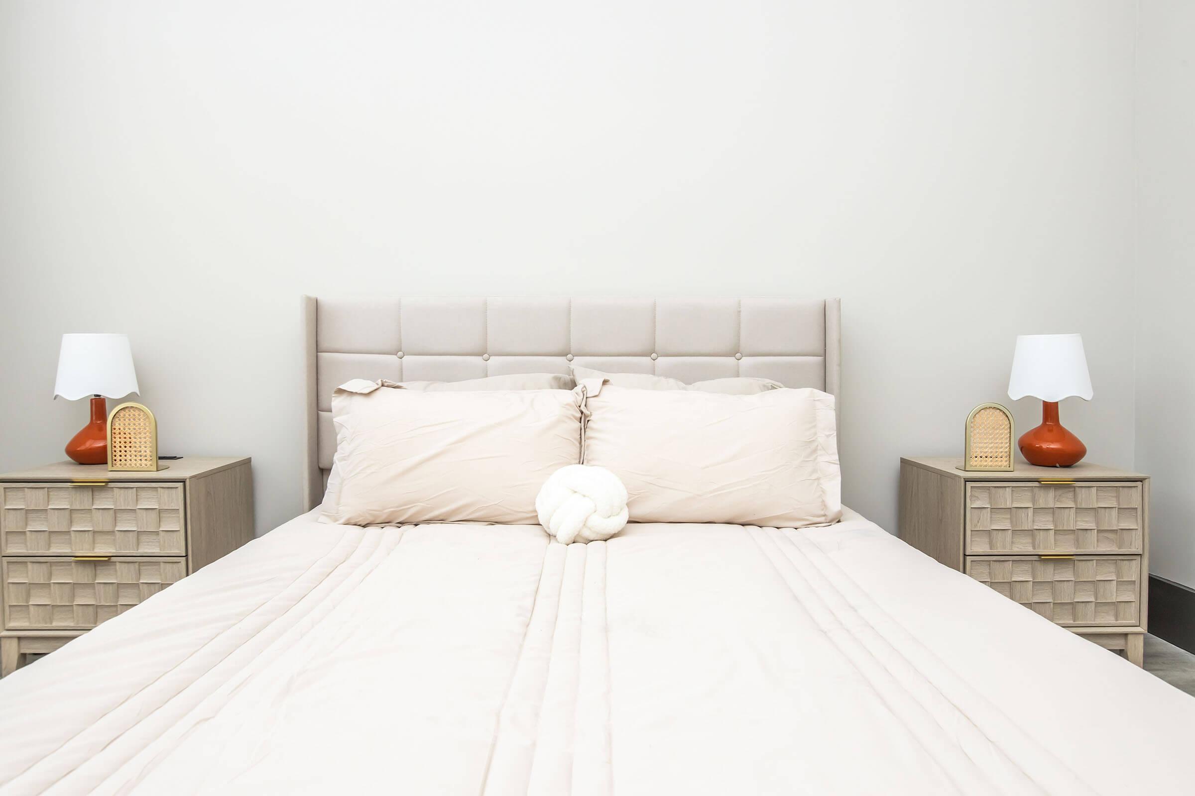 A light-colored bedroom featuring a queen-sized bed with a plush headboard. Two decorative pillows are arranged on the bed, along with a knotted throw pillow in the center. On each side, there are bedside tables with orange lamps and woven accents, against a neutral wall.