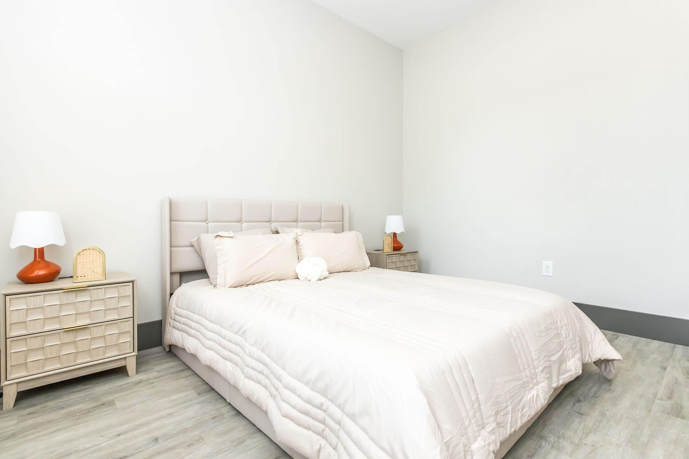 A softly lit bedroom featuring a light-colored bed with a beige comforter and decorative pillows. On either side of the bed, there are bedside tables with orange lamps. The walls are a soft neutral color, and the flooring is light-colored wood. The room has a minimalistic and modern design.