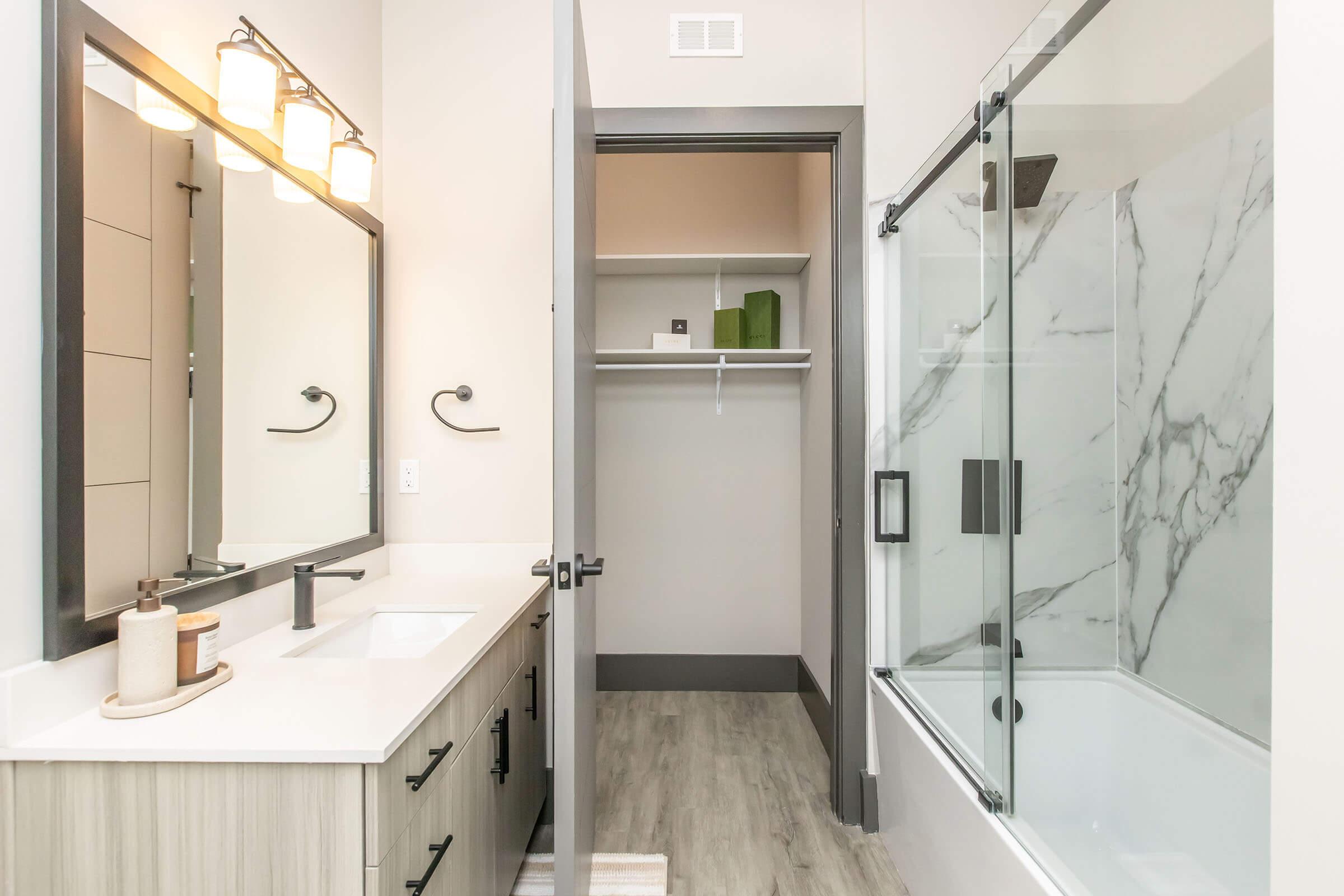 A modern bathroom featuring a double vanity with a large mirror, stylish light fixtures, and a glass-enclosed shower with marble accents. A door leads to a closet area with shelves, and there are neutral-colored walls and flooring for a clean, contemporary look.