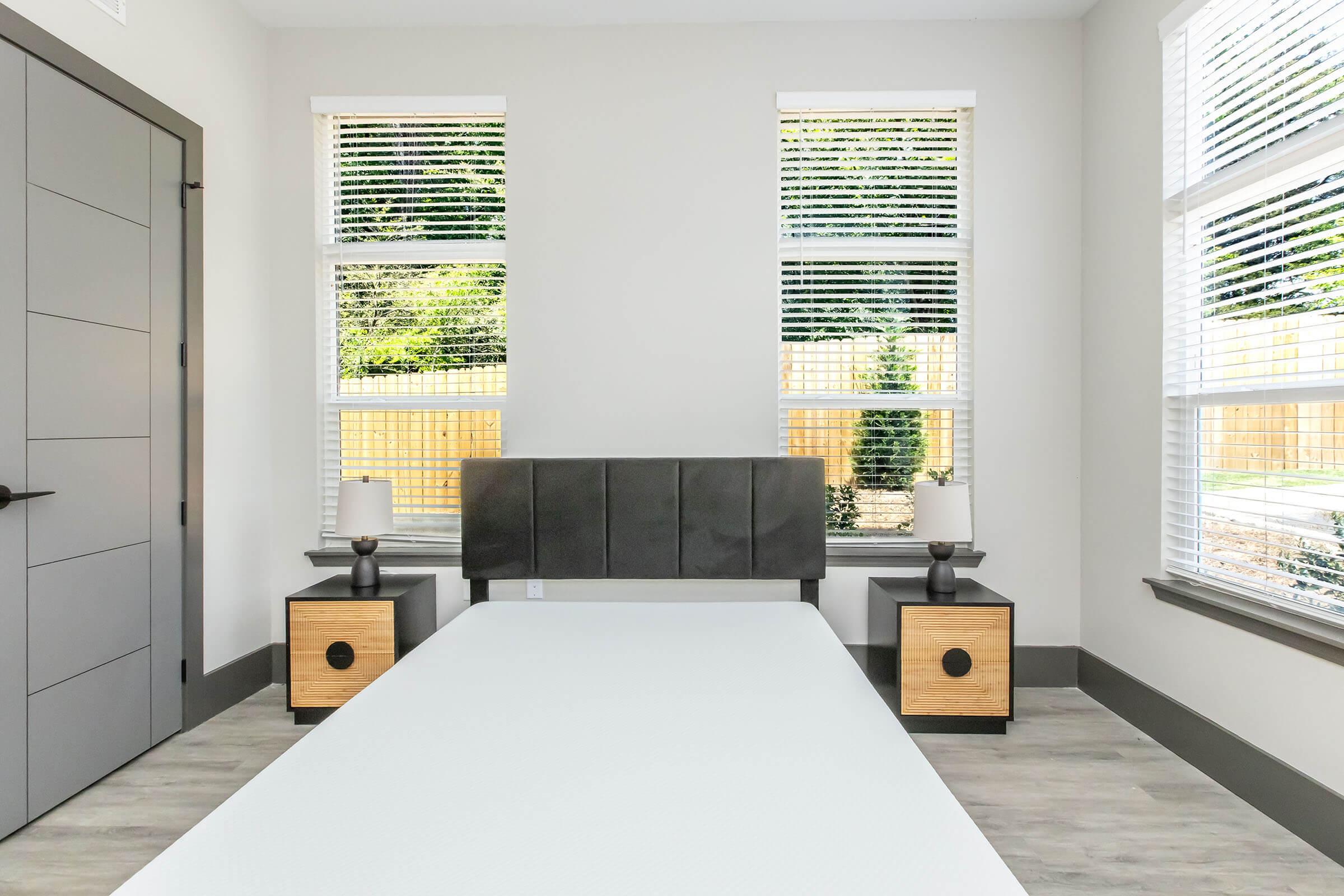 A minimalist bedroom featuring a simple bed frame with a white mattress, flanked by two modern nightstands with lamps. The room has large windows with blinds, allowing natural light to enter. Light-colored walls and wood flooring create a bright and airy atmosphere.