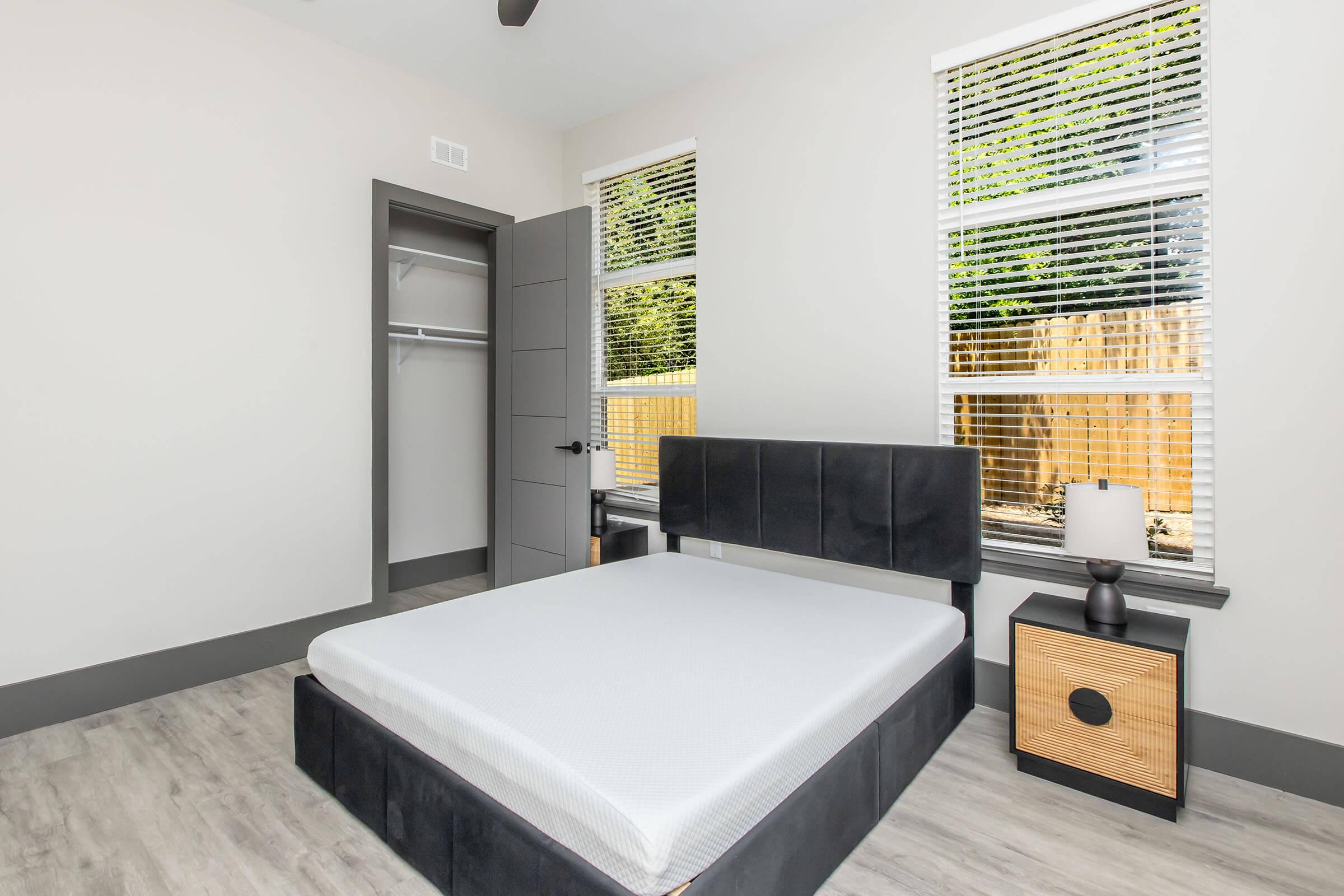 A modern bedroom featuring a black bed frame with a white mattress, a nightstand with a lamp, and large windows with white blinds. The walls are a light color, and there is a closet with an open door. The flooring is light wood, and a wooden fence is visible outside the windows.