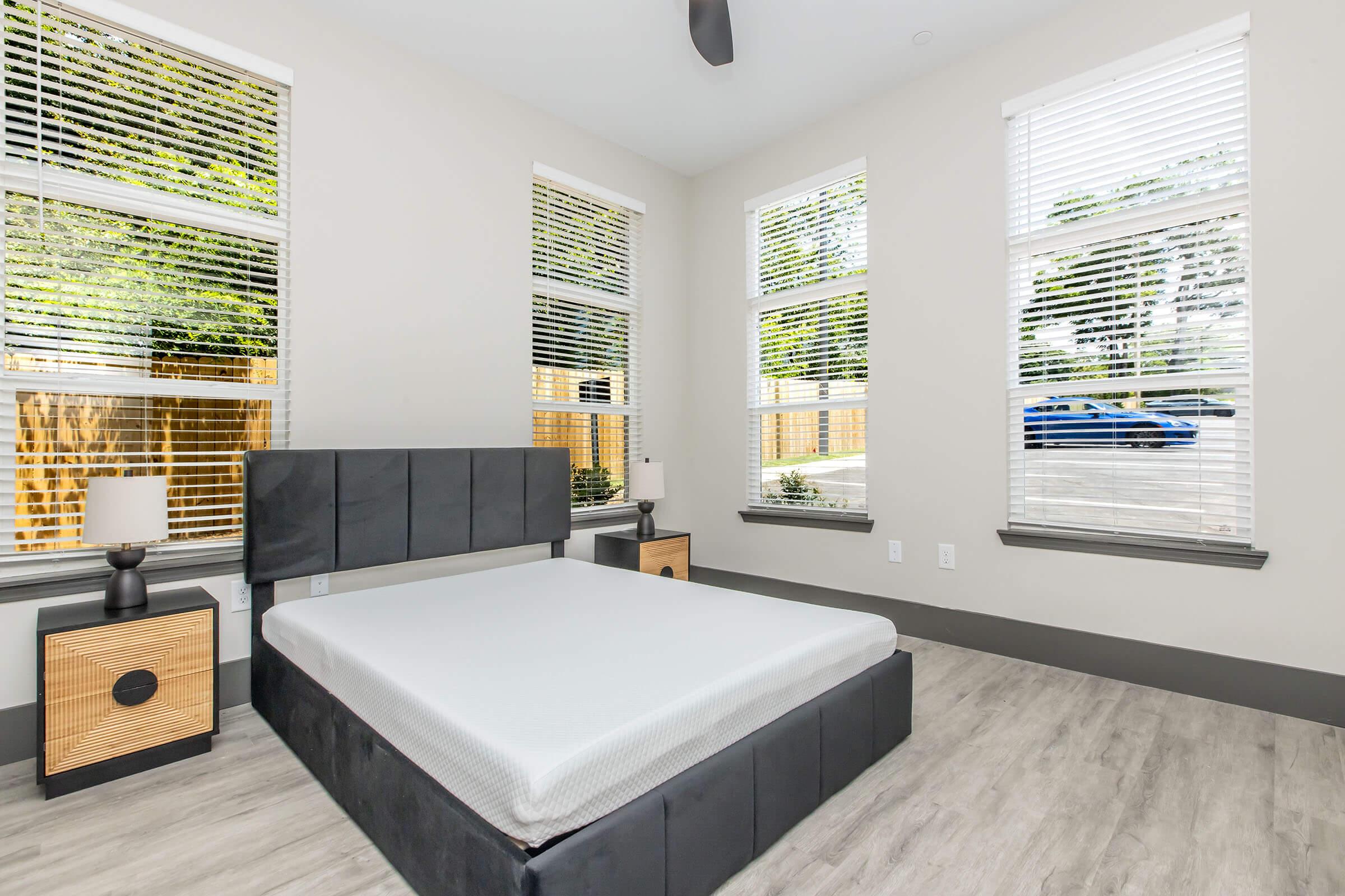 A modern bedroom featuring a gray platform bed with no mattress, two bedside tables with lamps, large windows with white blinds, and light-colored wooden flooring. Natural light illuminates the space, highlighting the contemporary design and minimalistic decor.