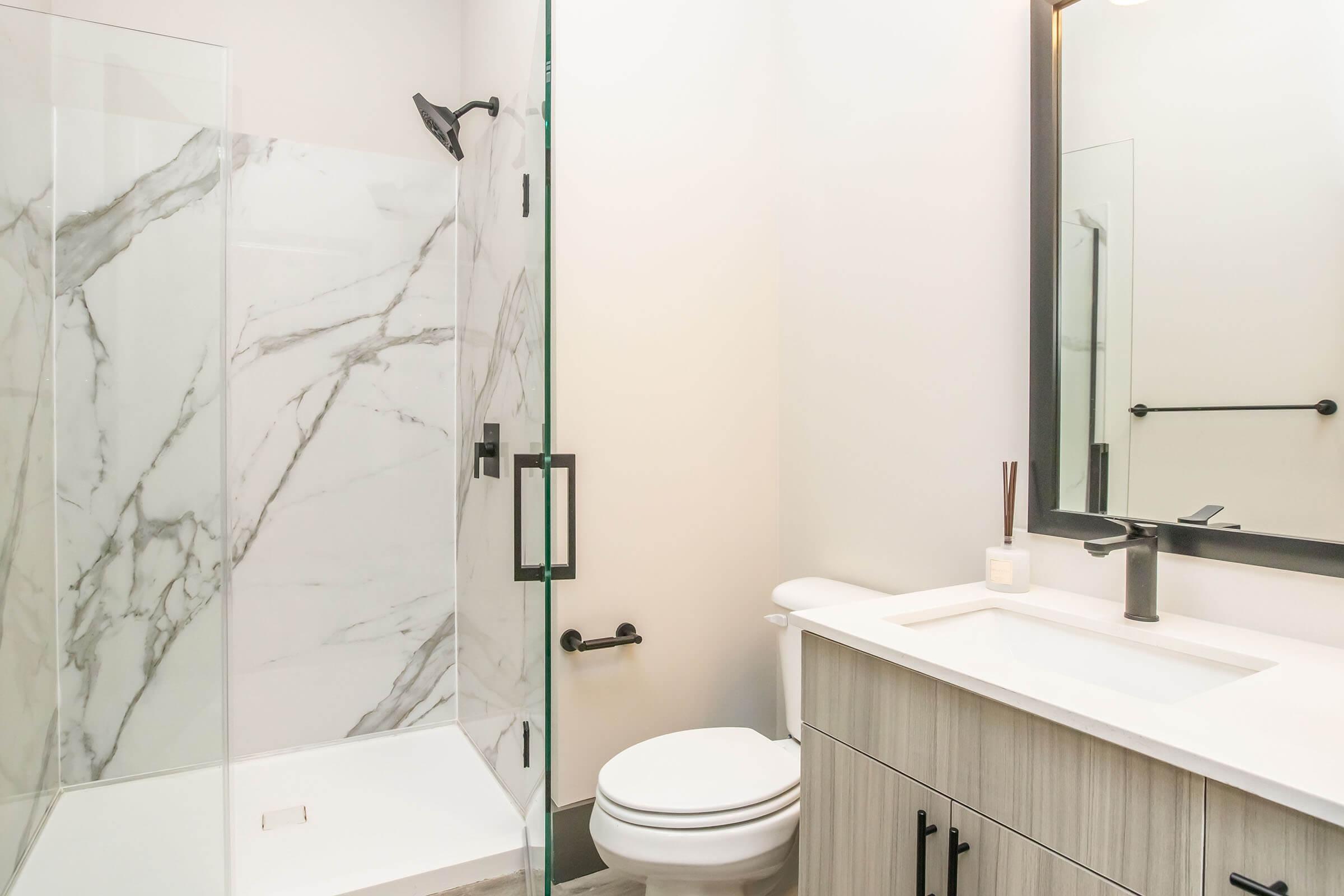 Modern bathroom featuring a walk-in shower with marble-patterned walls, a white toilet, and a sleek vanity with a minimalist design. The space is well-lit, with neutral colors and contemporary fixtures, creating a clean and stylish atmosphere.