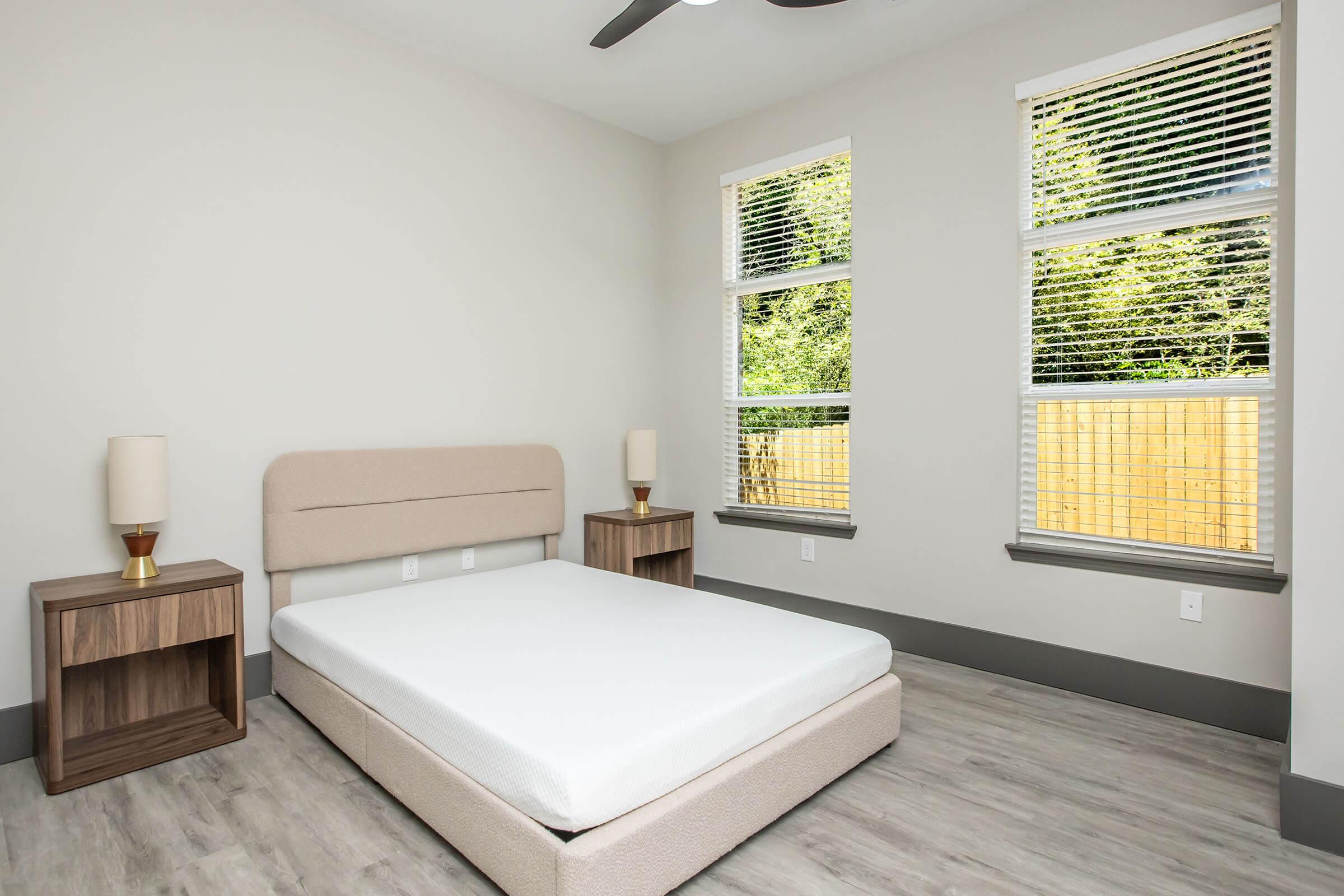 A minimalist bedroom featuring a beige bed frame without a mattress, wooden nightstands with lamps on either side, and large windows with white blinds letting in natural light. The walls are painted light gray, and there is a light wooden floor.