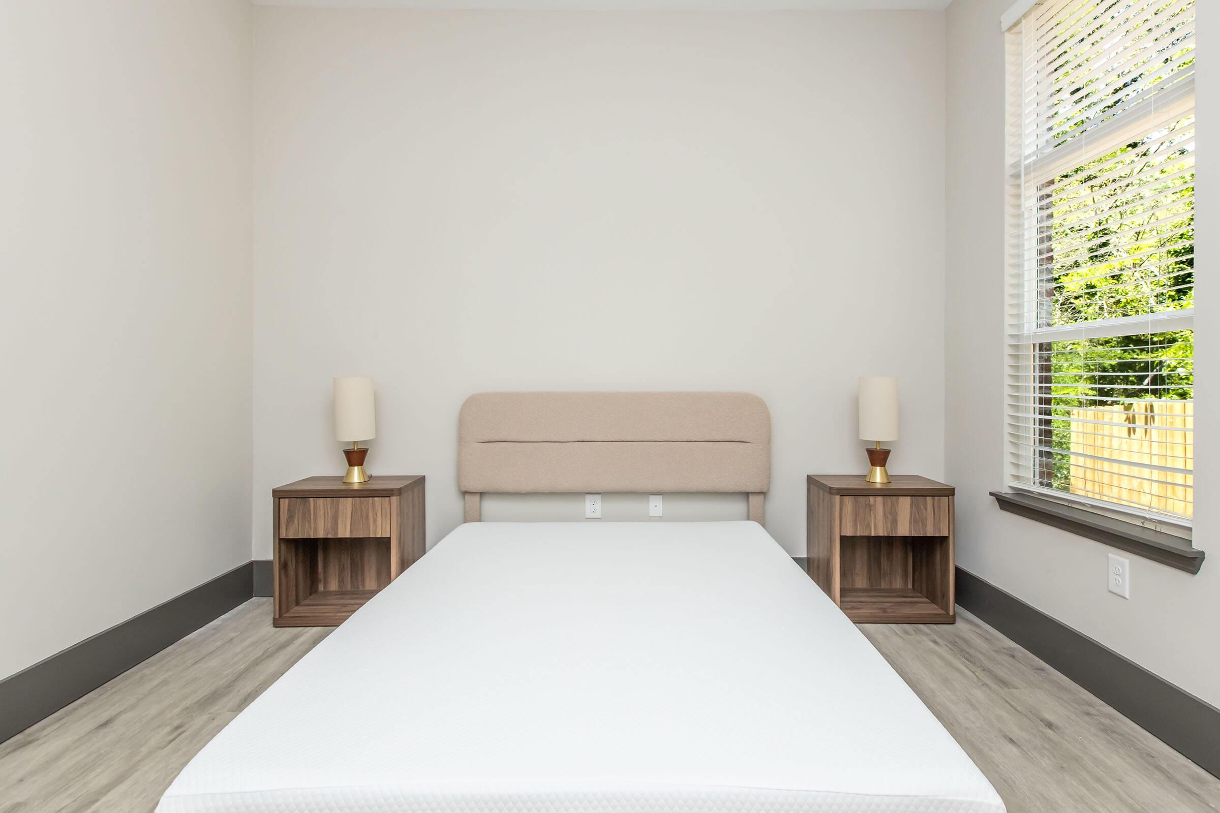 A minimalistic bedroom featuring a light-colored wall, a queen-sized bed with a beige upholstered headboard, and two wooden nightstands with lamps. The floor is light wood, and a window allows natural light to enter, revealing greenery outside.