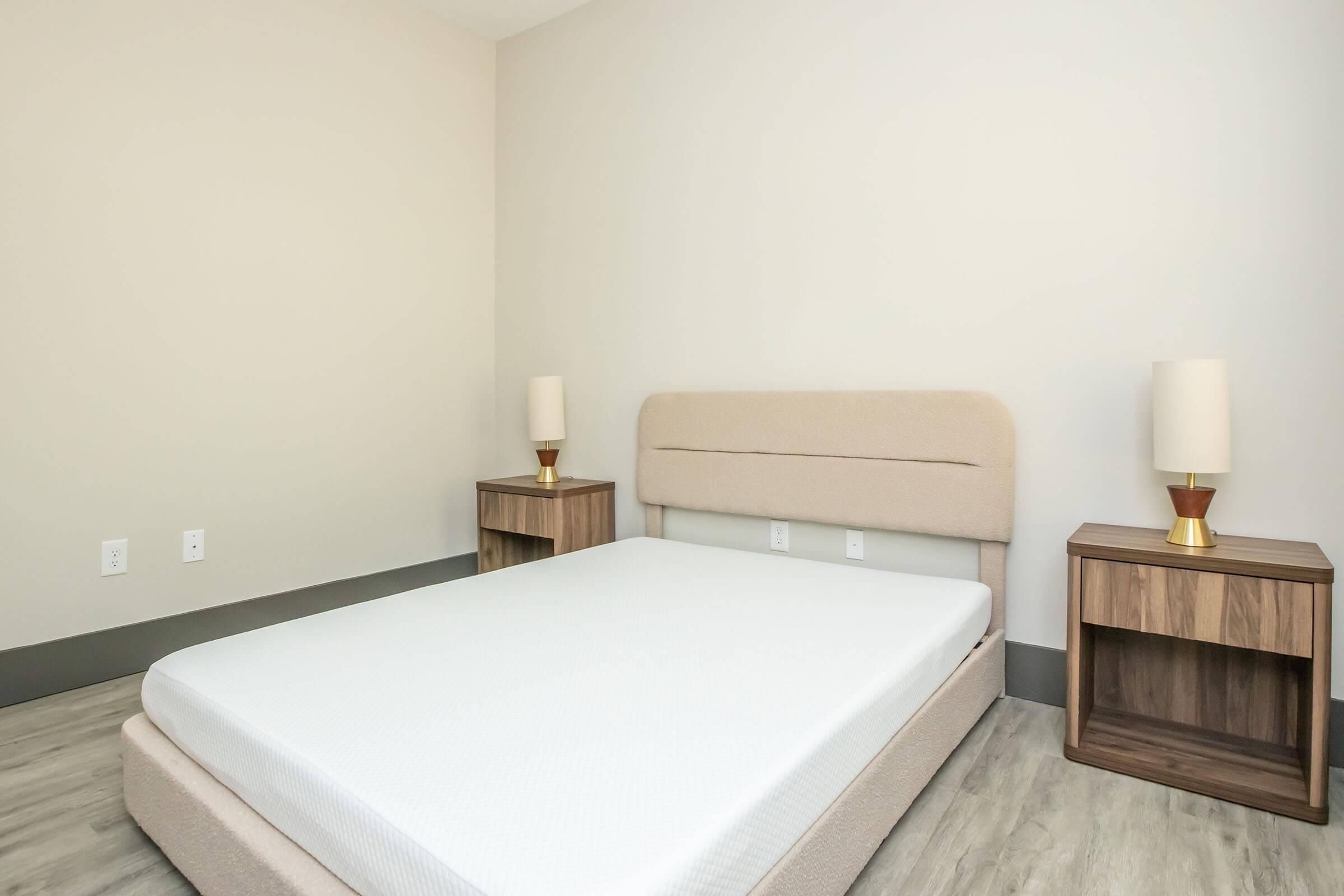 A minimalistic, empty bedroom featuring a light-colored upholstered bed frame without bedding, two wooden nightstands with neutral-colored lamps on each side, and a simple, clean wall. The flooring is light-colored wood, and the overall aesthetic is modern and uncluttered.