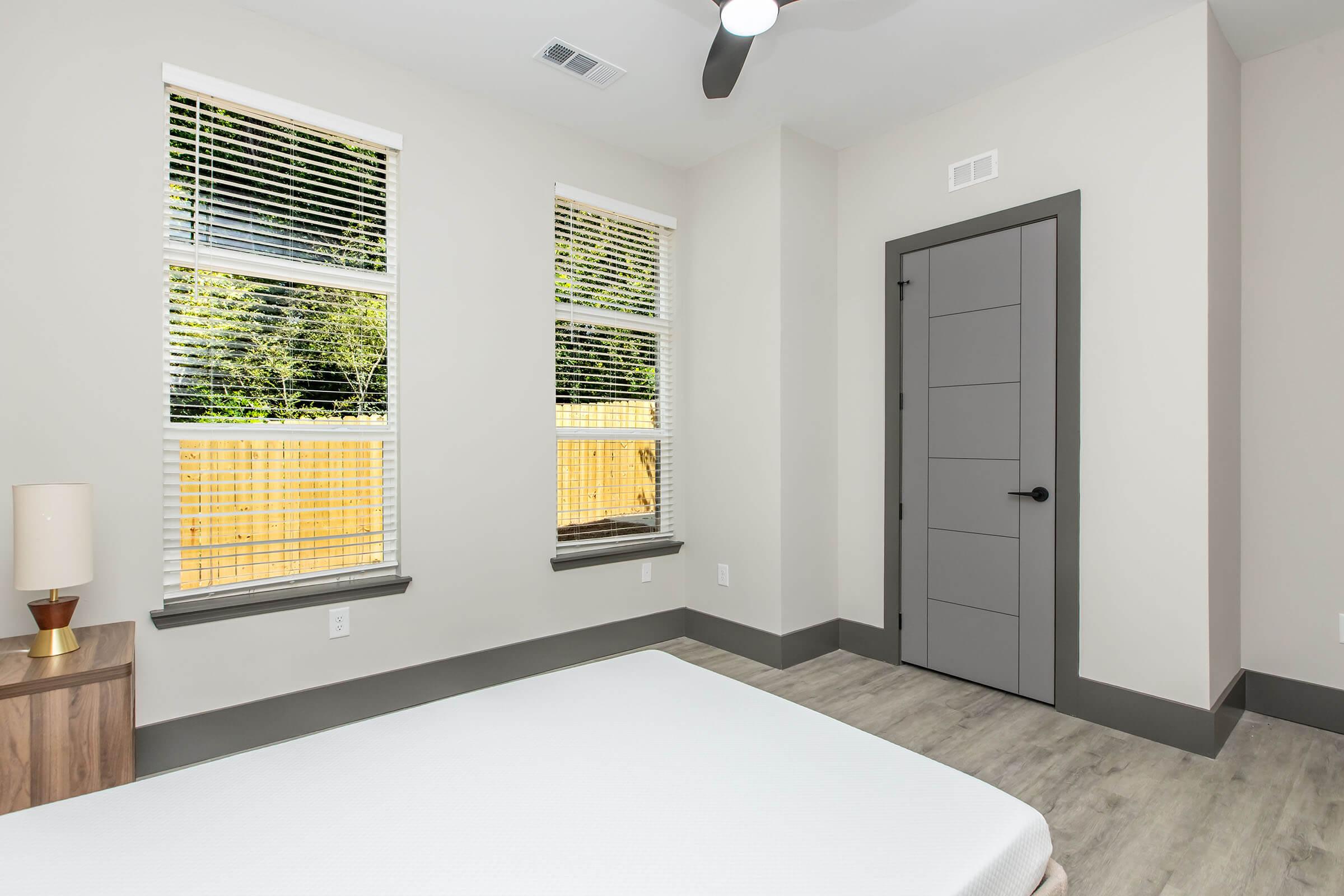 A modern, empty bedroom featuring light-colored walls and large windows with blinds. There's a wooden nightstand with a lamp beside a bare mattress on the floor. The door has a sleek design, and the room has a clean, minimalist aesthetic with natural light illuminating the space.
