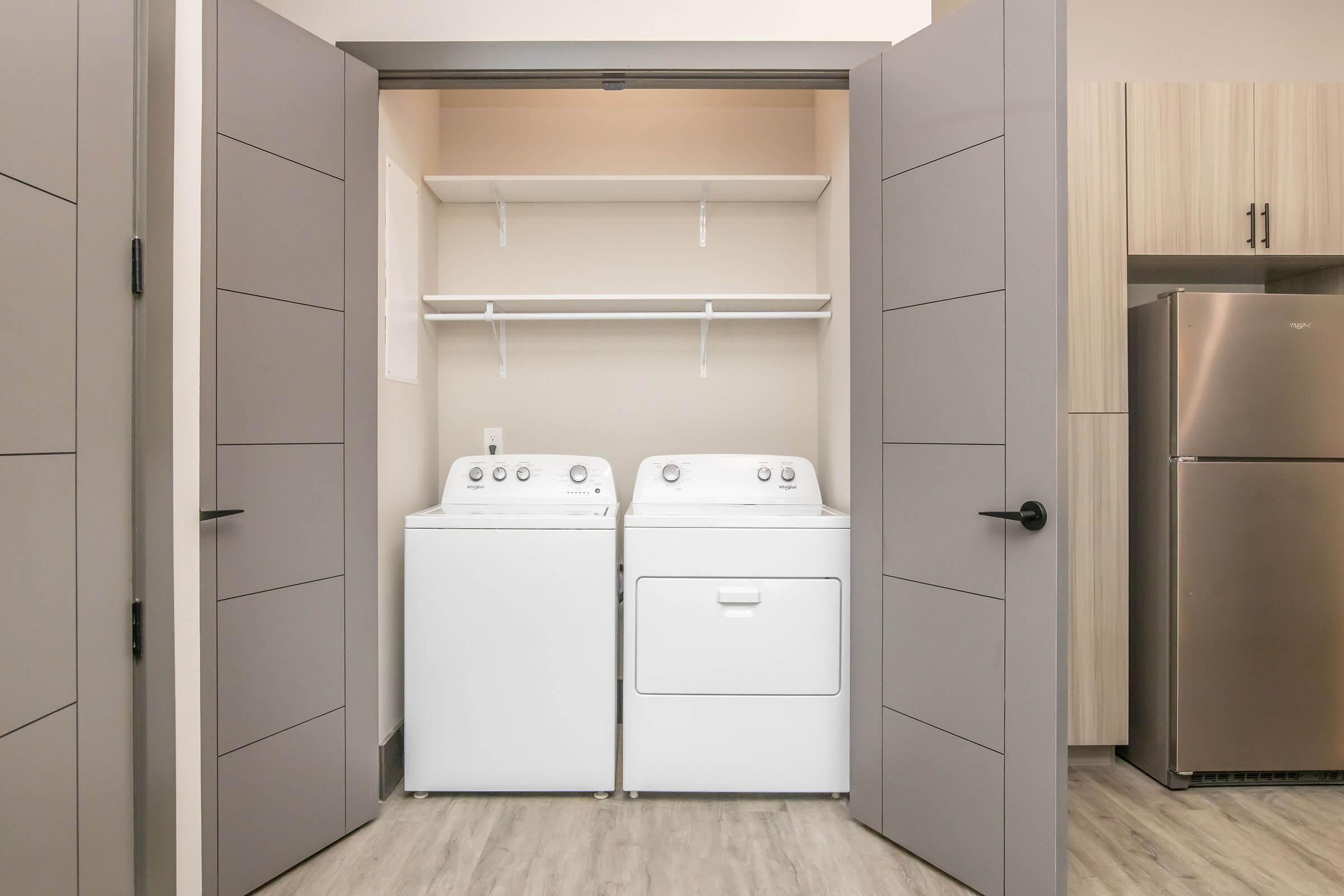 A laundry closet featuring a stacked washer and dryer set, with a shelf above for storage. The closet has modern gray doors, and there is a stainless steel refrigerator visible to the right, set within a contemporary kitchen environment. The flooring is light-colored and sleek.