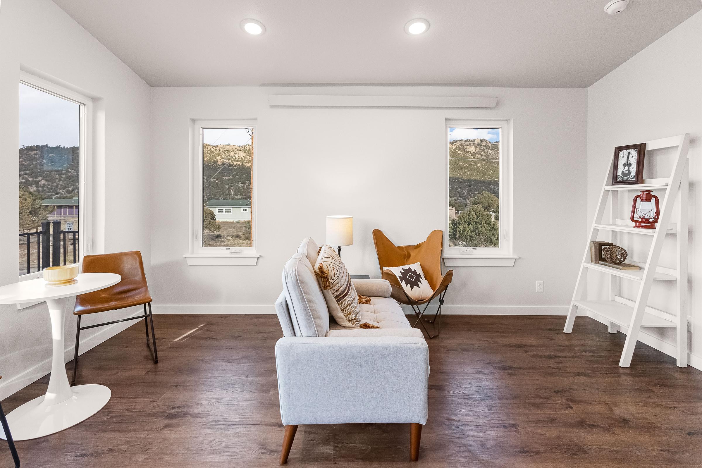 A cozy living room featuring a light gray sofa with decorative pillows, a white round dining table with a chair, and a stylish orange butterfly chair. Large windows provide a view of the outdoors, and a white bookshelf displays decorative items against a neutral wall. Modern and inviting atmosphere.