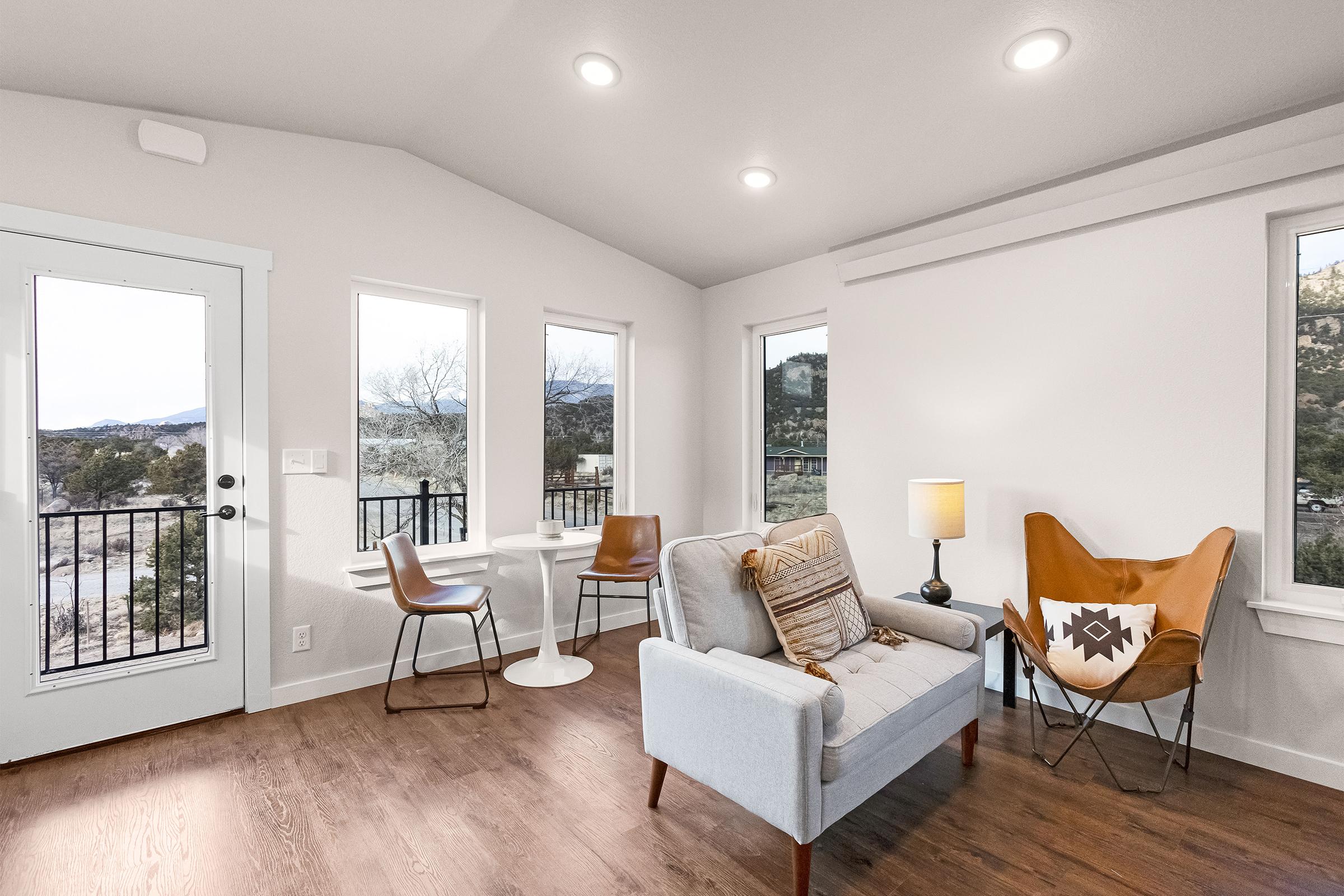 A modern living room featuring a light gray sofa with decorative pillows, a unique butterfly chair, a small white side table, and two wooden chairs. Large windows allow natural light and offer a view of the outdoors. The space has a warm wooden floor and minimalist decor.