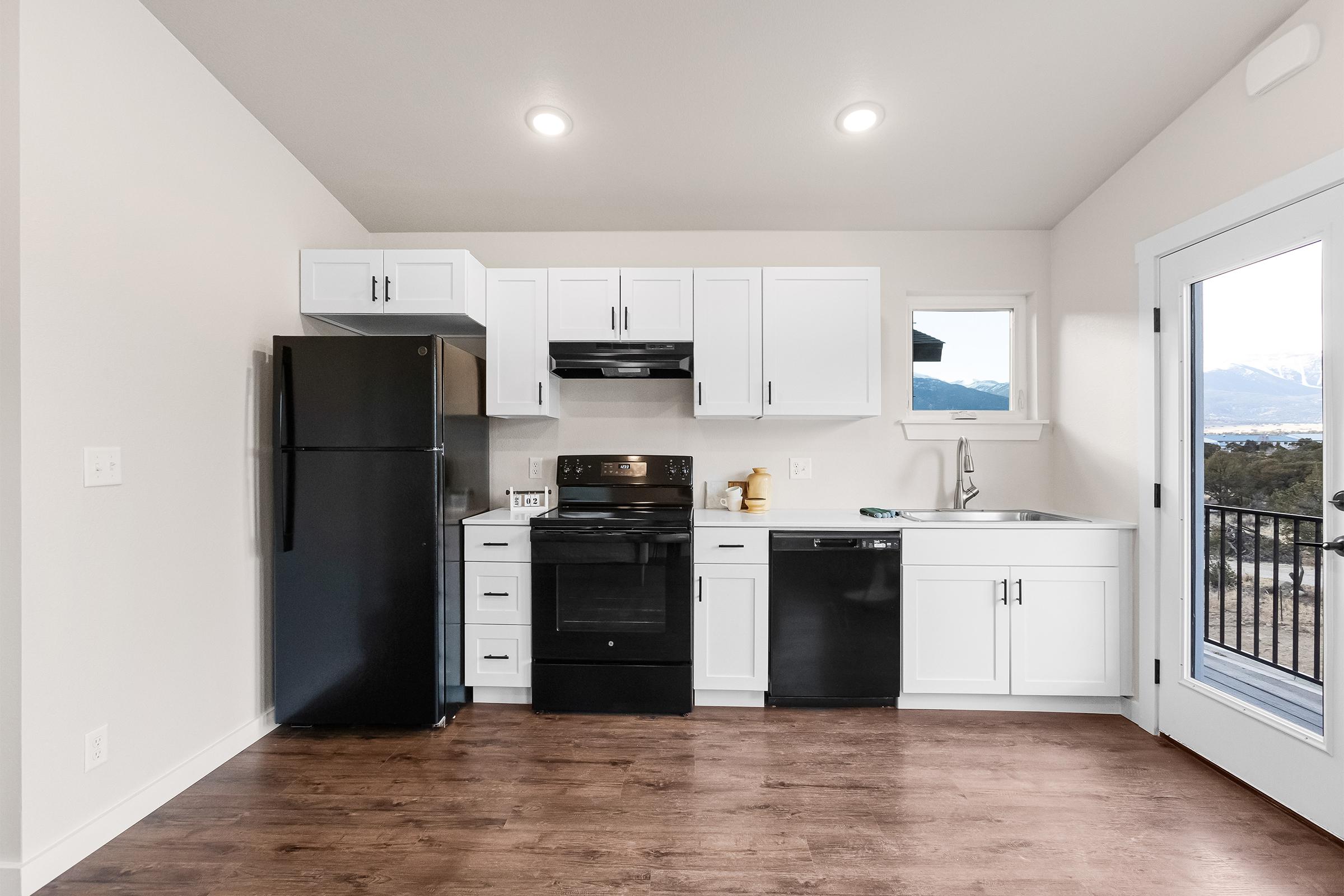 A modern kitchen featuring white cabinets, black appliances including a refrigerator, stove, and dishwasher. There's a window showcasing a view, and the flooring is a warm wood tone, creating a bright and inviting atmosphere.