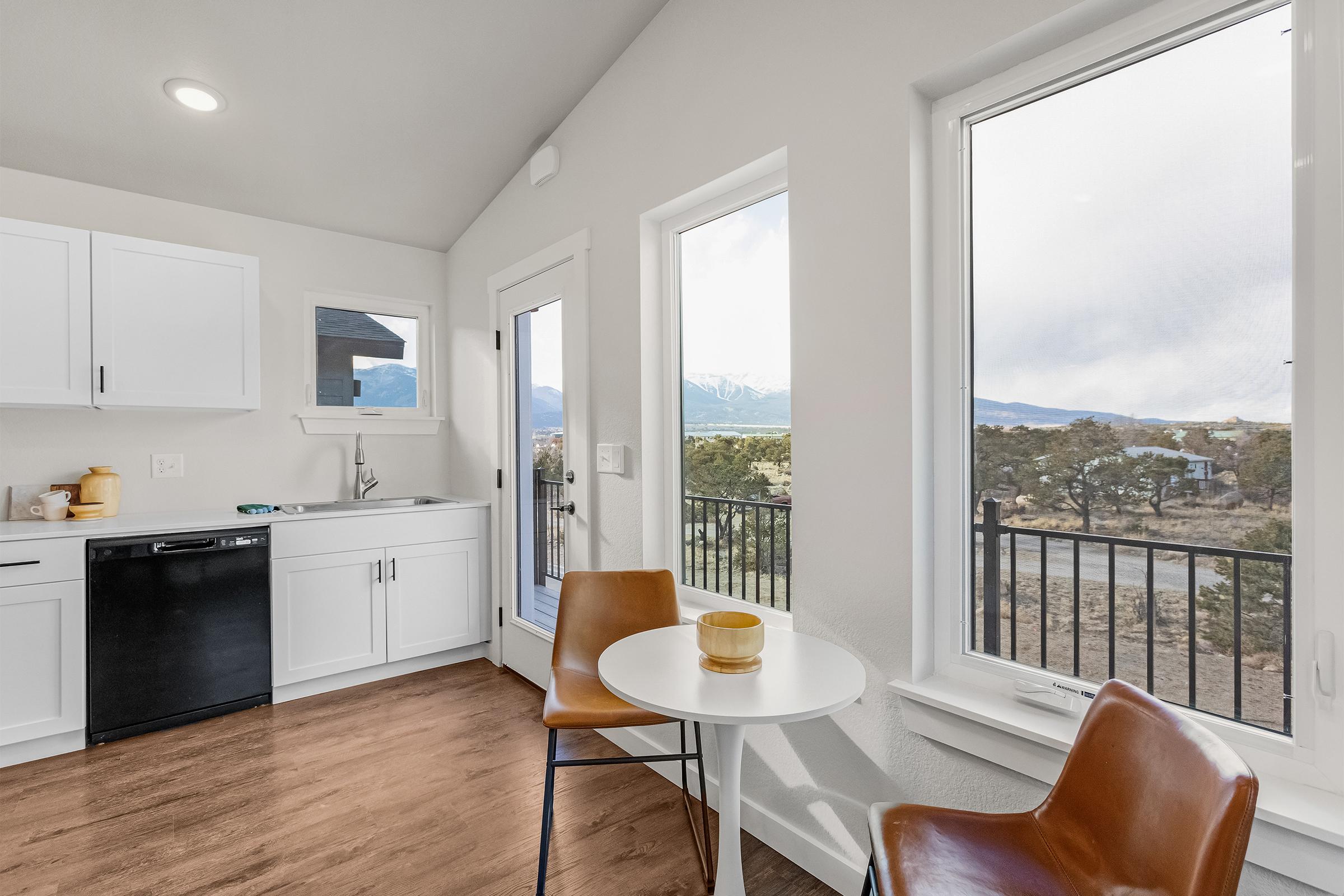 A modern kitchen area featuring white cabinets, a small round table with two chairs, and large windows showcasing a scenic mountain view. The kitchen includes a sink and black dishwasher, and the flooring is a warm wood tone. Bright natural light illuminates the space.