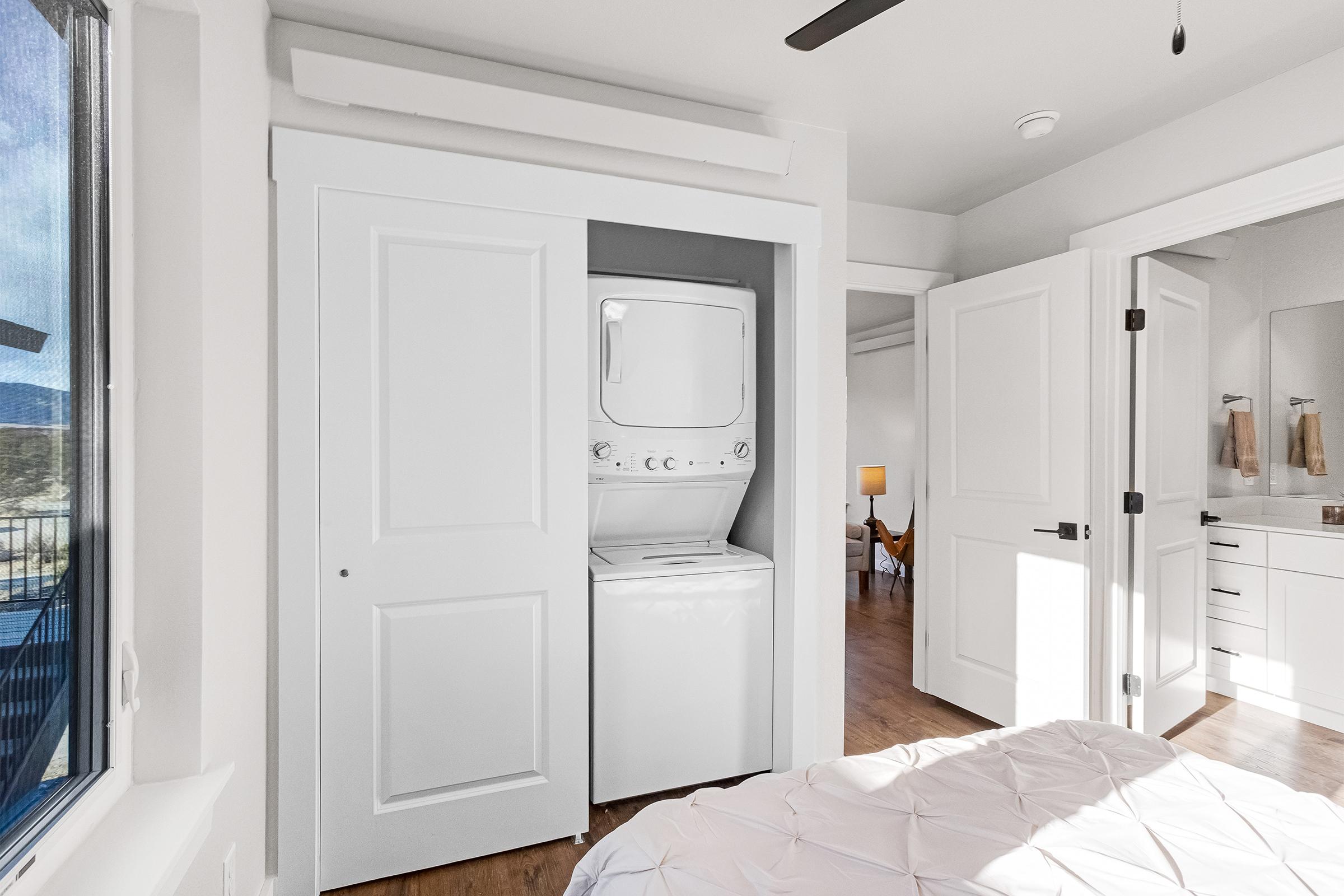 A compact laundry area featuring a stacked washer and dryer, enclosed within double white doors. The room has modern finishes and hardwood flooring, with natural light coming from a nearby window. The space is neatly organized, suggesting a contemporary living environment.