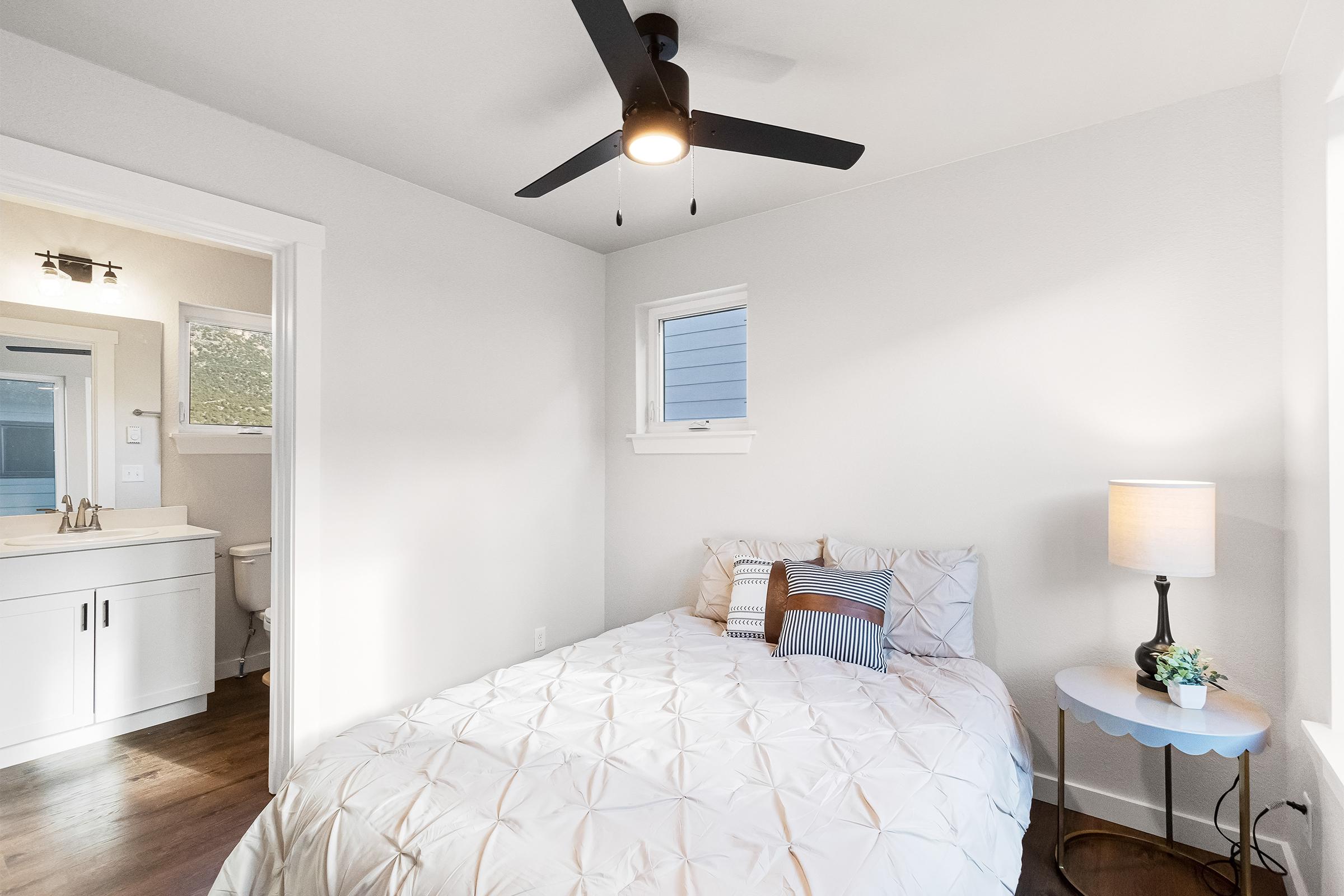 A bright and cozy bedroom featuring a queen-sized bed with decorative pillows, a bedside lamp on a small table, and a ceiling fan. A bathroom is visible through an open door, with natural light coming in from a window. The walls are painted white, creating a clean and modern look.